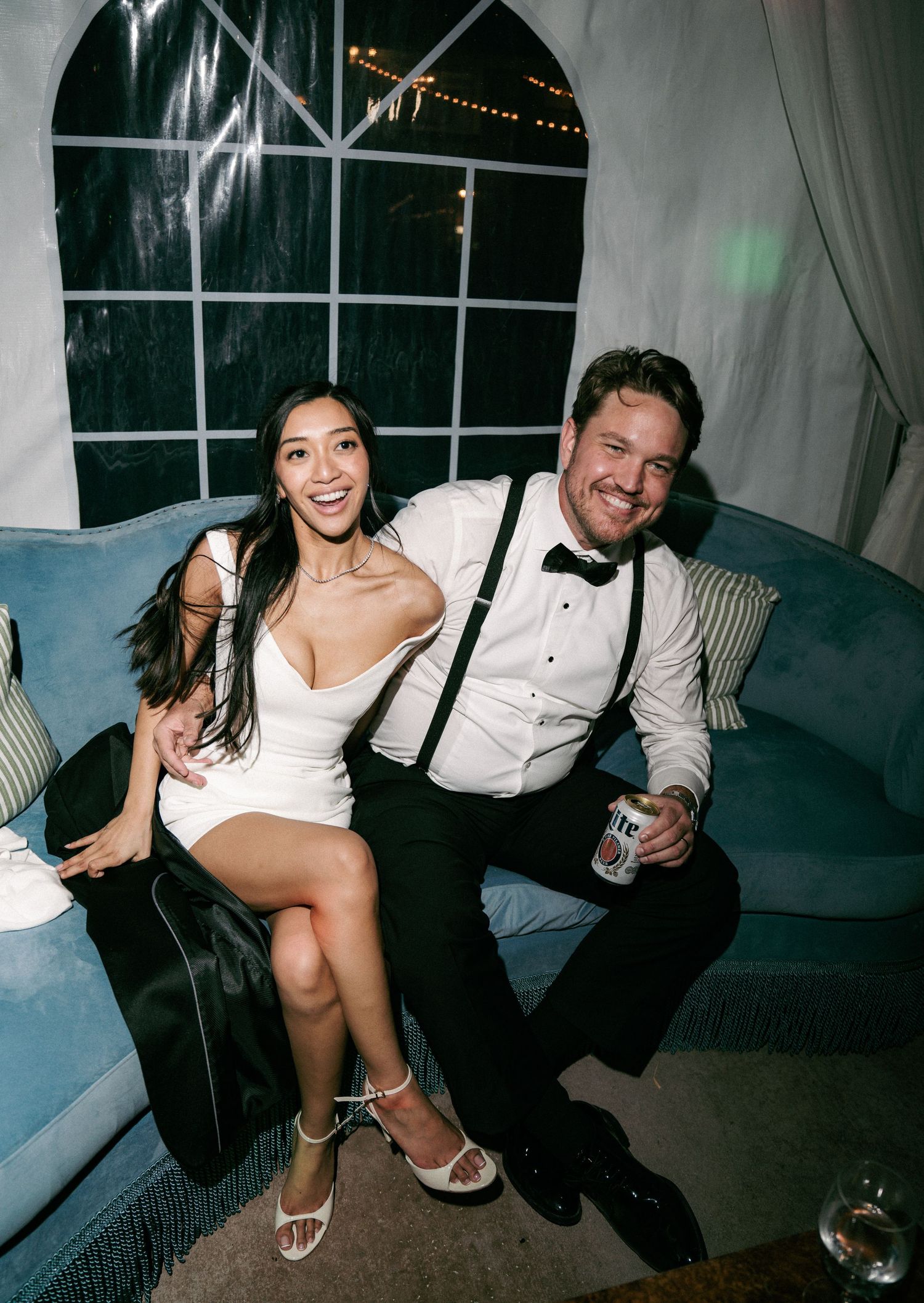Couple in formal attire smiling on a blue sofa at an elegant event, woman in white dress, man in tuxedo with suspenders.