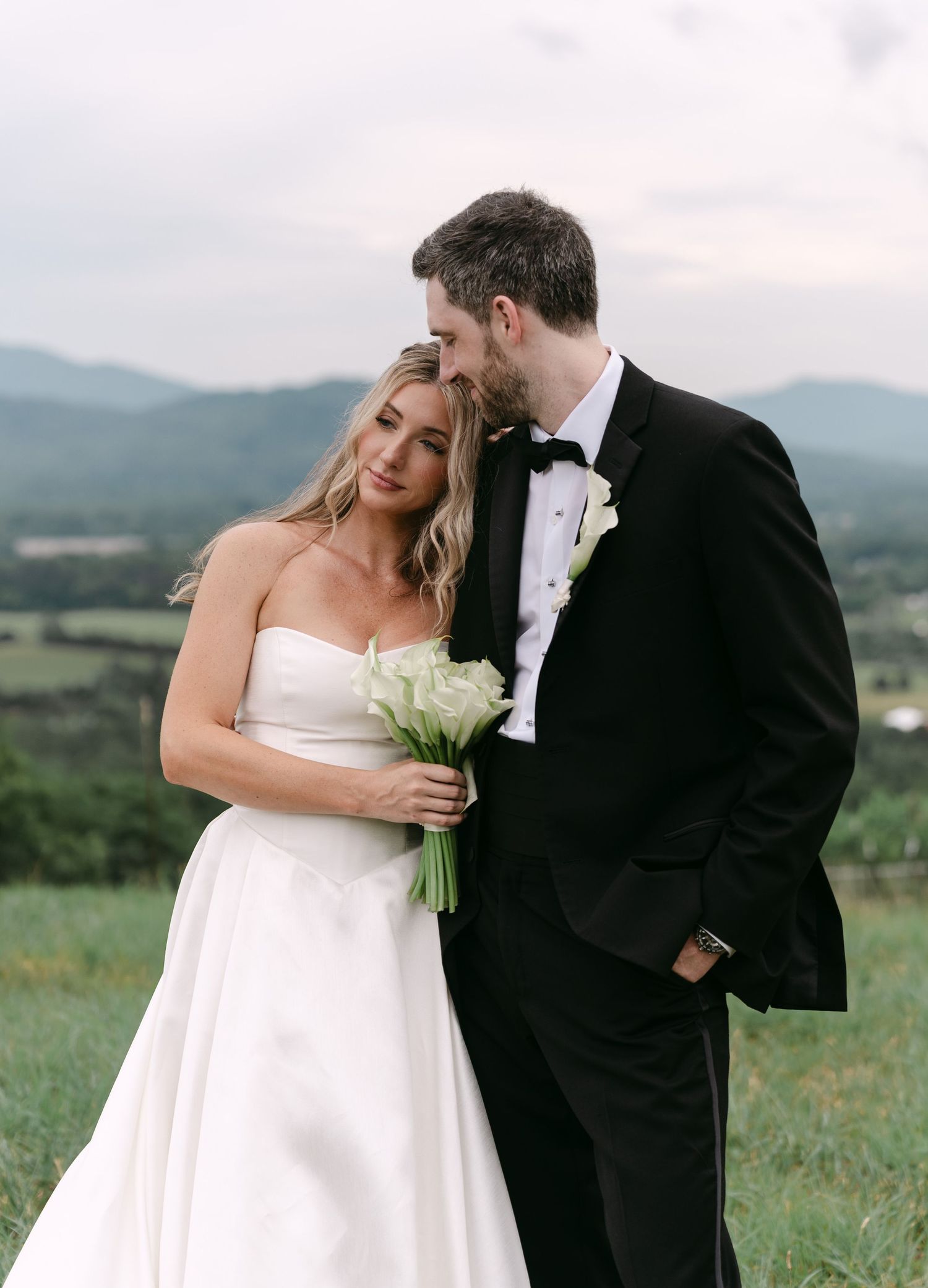 Bride in strapless white gown holding white bouquet leans into groom in black tuxedo with mountain backdrop.