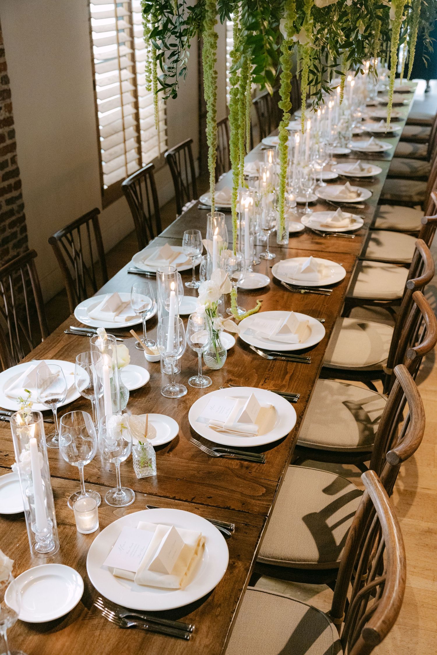 Long wooden dining table set for elegant wedding reception with white place settings and tall glass centerpieces.