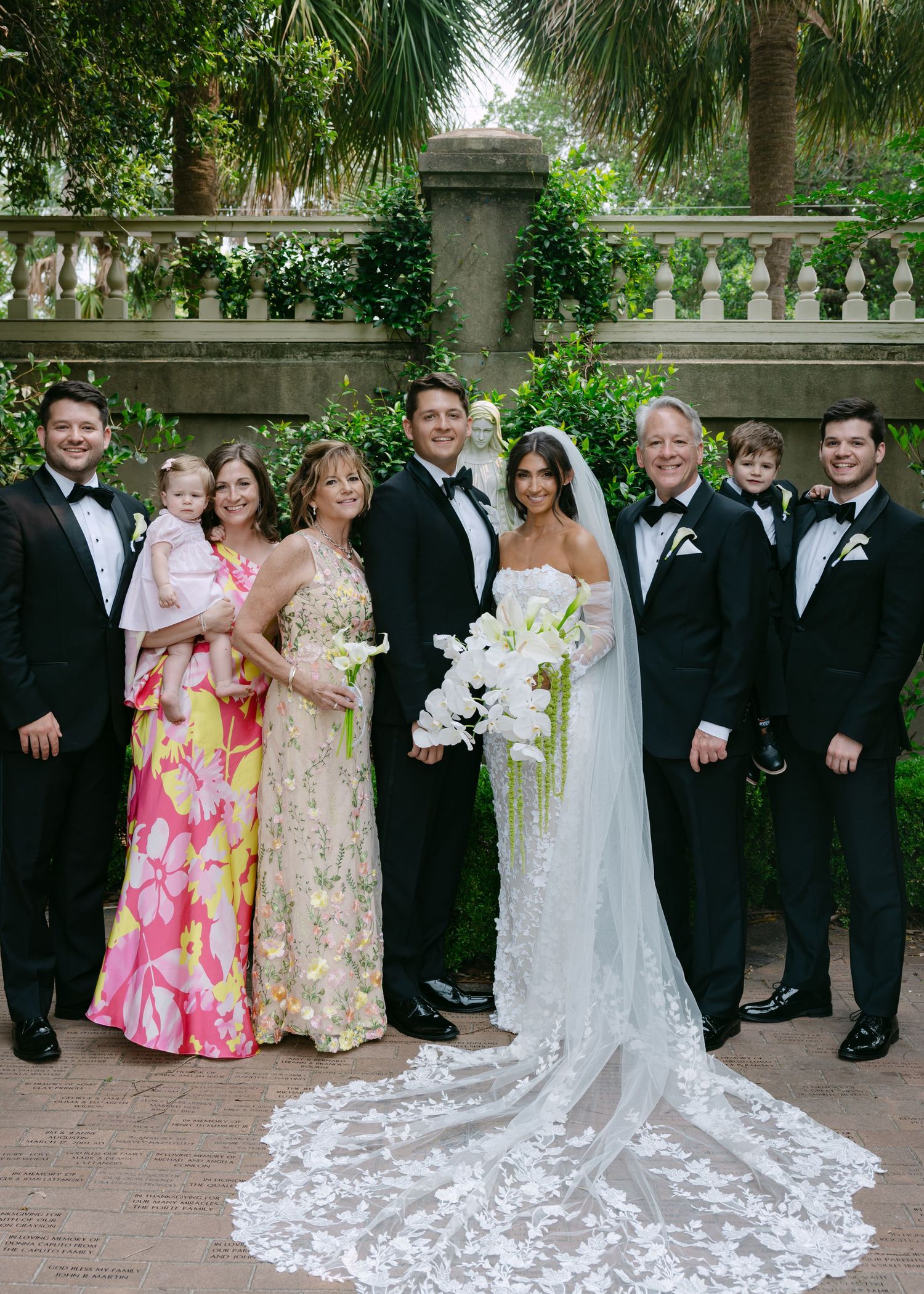 Wedding party members gathered for formal photos in a lush garden setting with classic architecture.