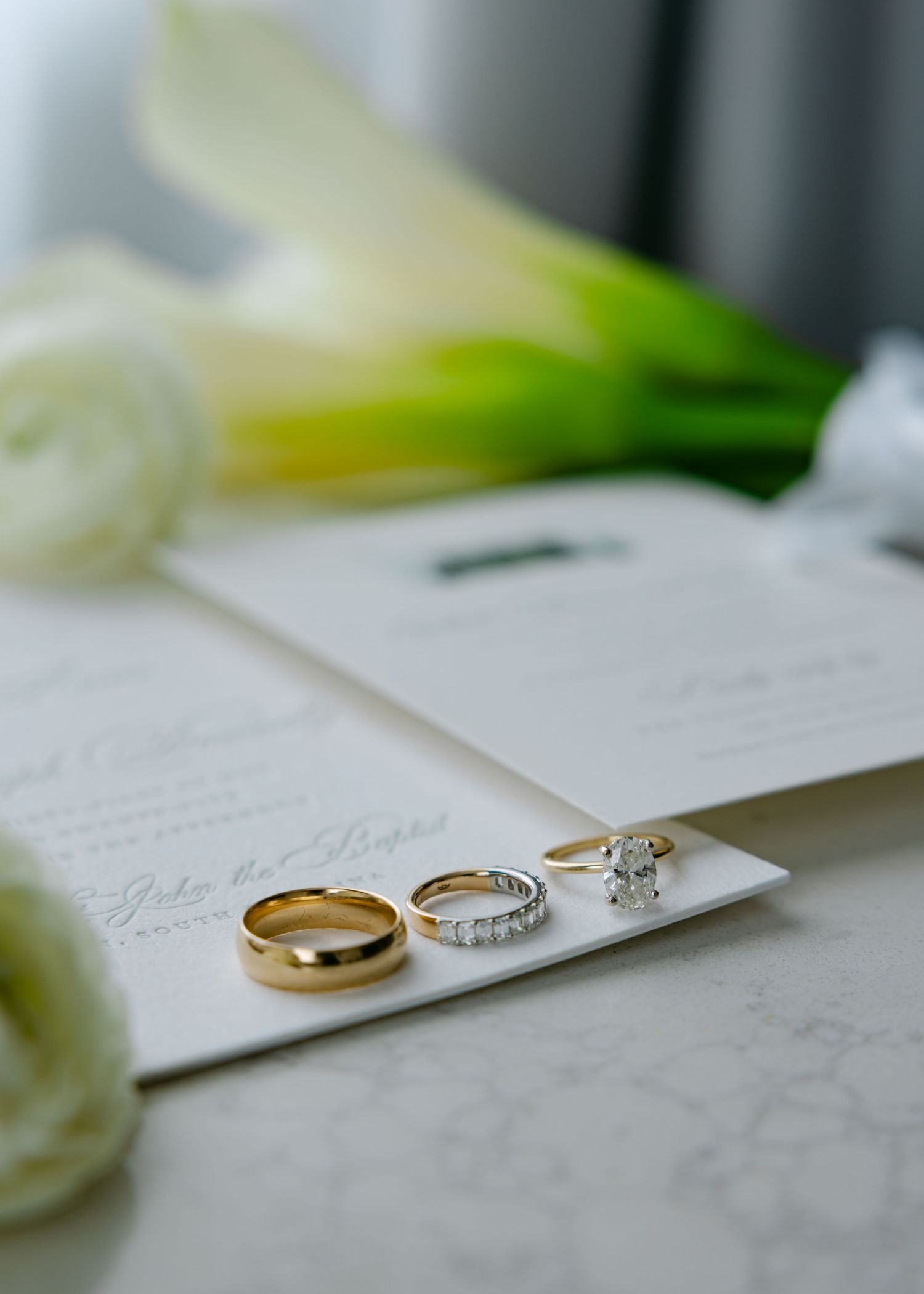 Wedding bands and engagement ring arranged on white invitation with calla lilies.