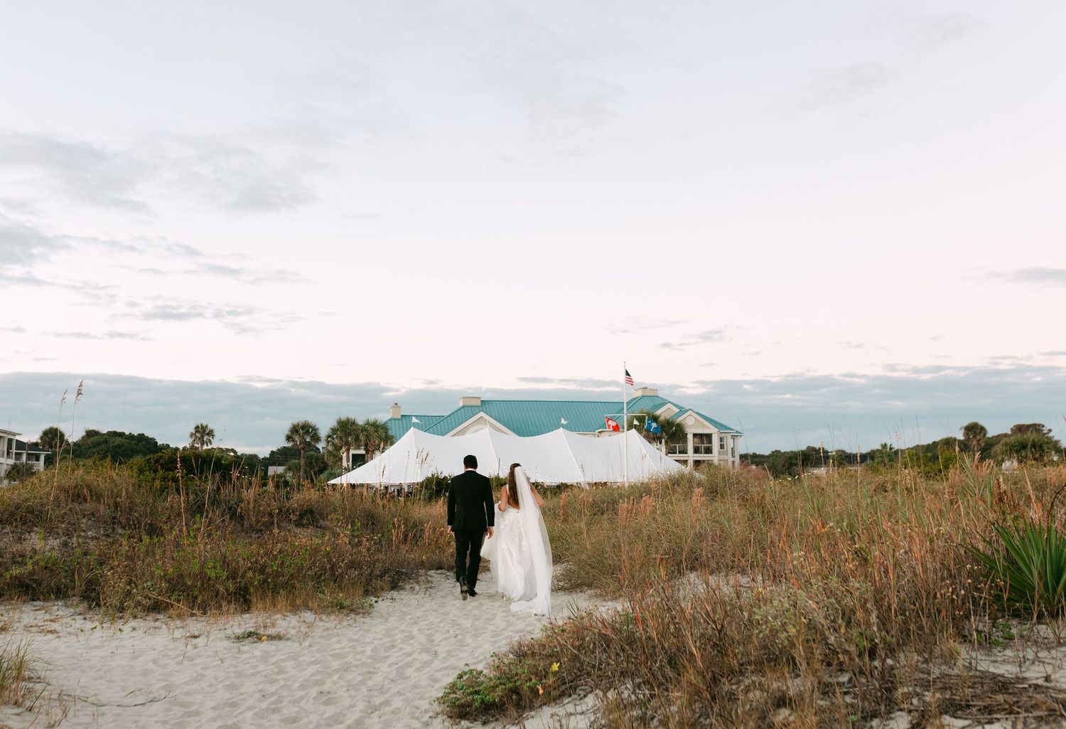 Bride and groom walk hand in hand toward a white sperry tent on a sandy beach path.
