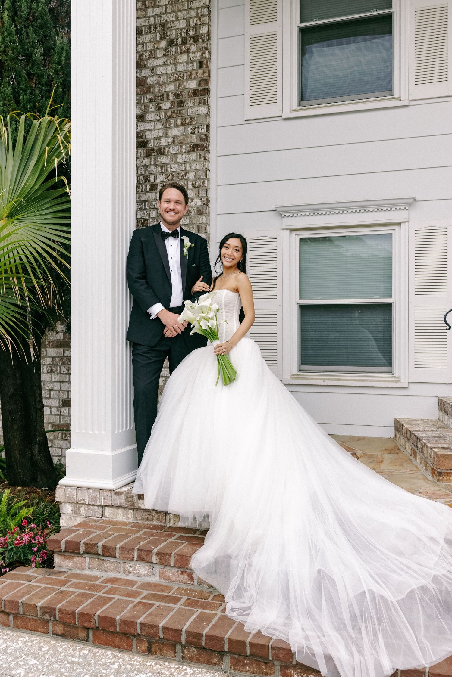 Smiling bride in strapless ballgown with groom in tuxedo posing on brick steps holding calla lilies.