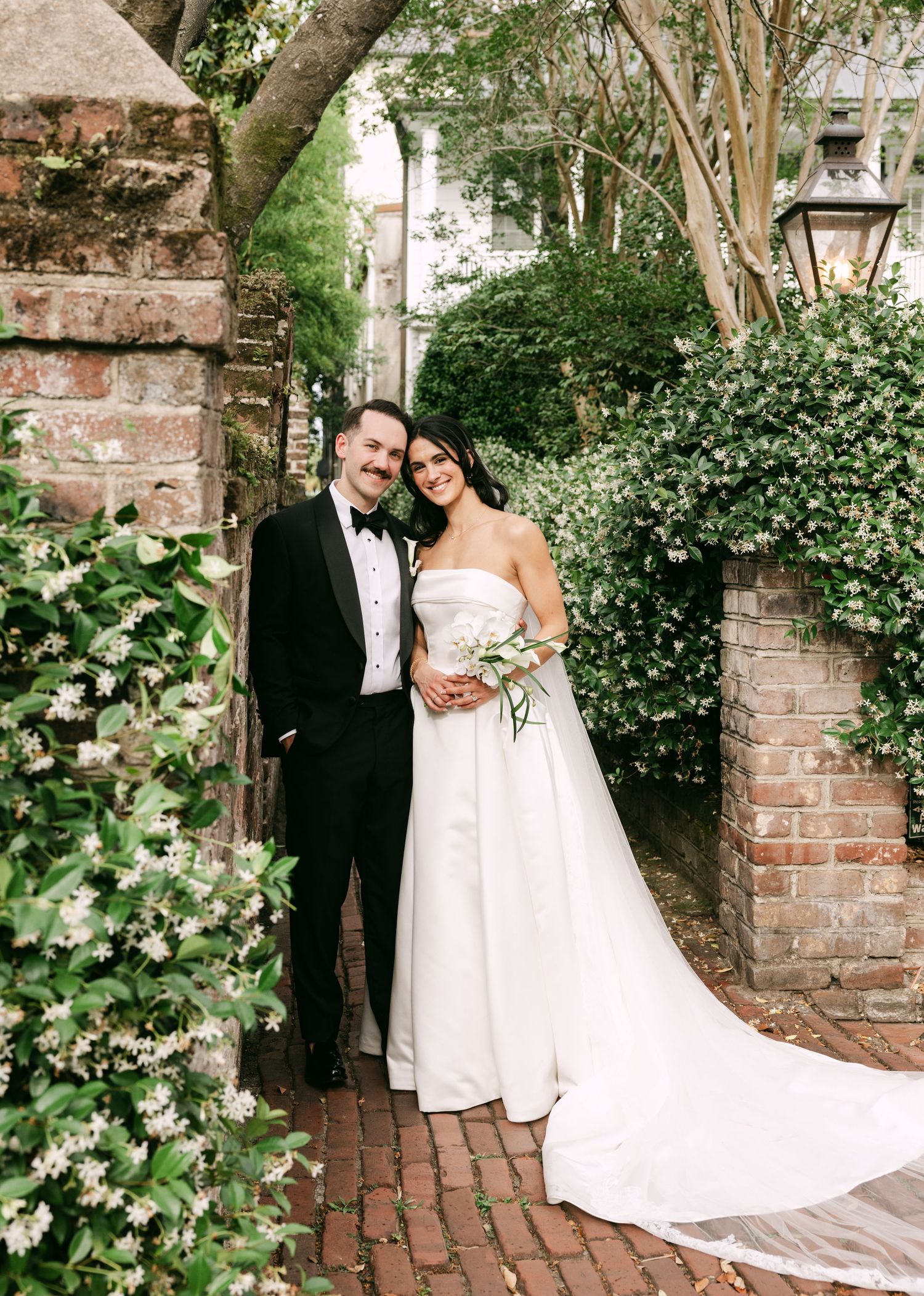 Bride and groom posing under a brick archway surrounded by lush green ivy and white flowers.