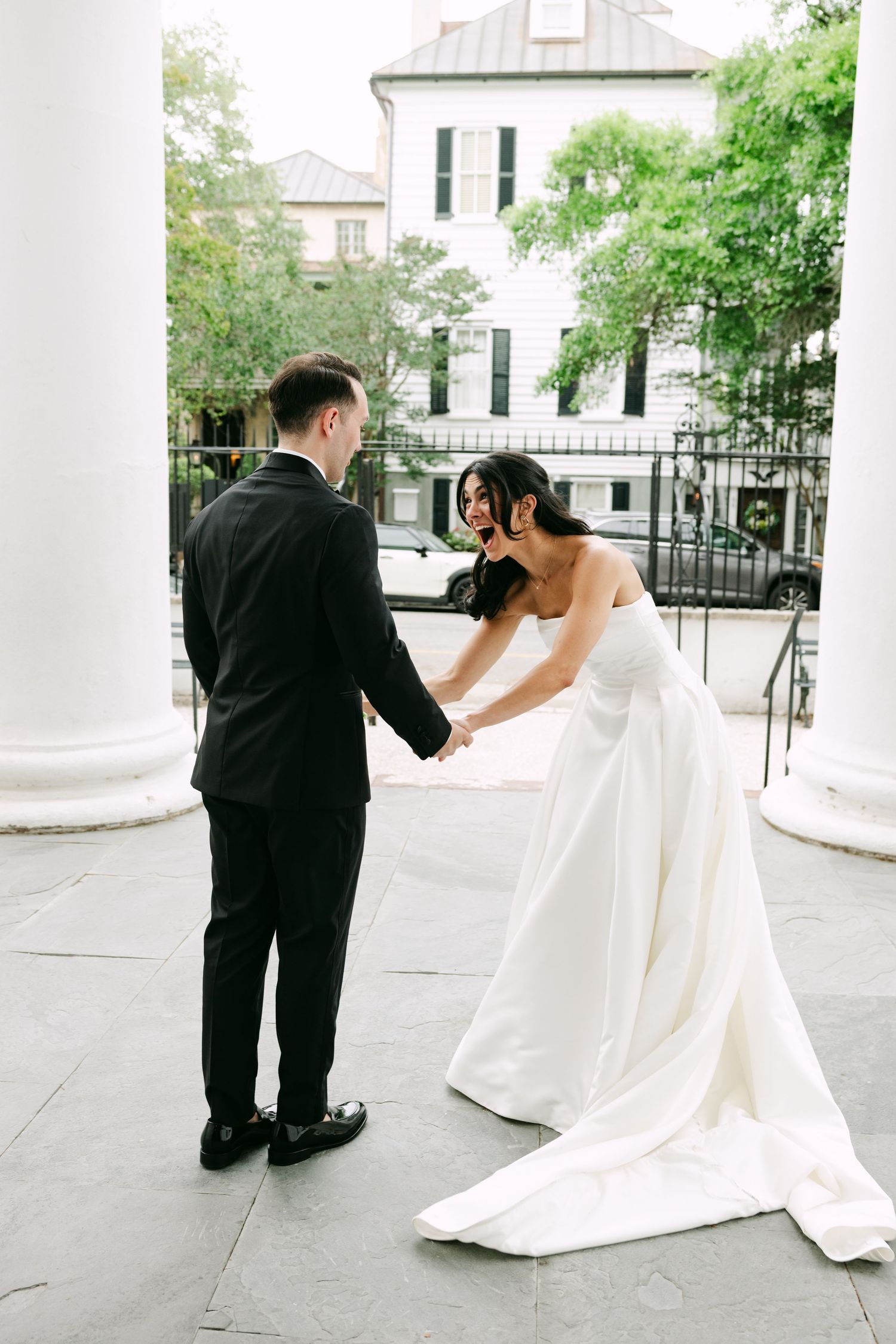 Bride in strapless white gown holds groom's hands during first look on stone steps with white columns.