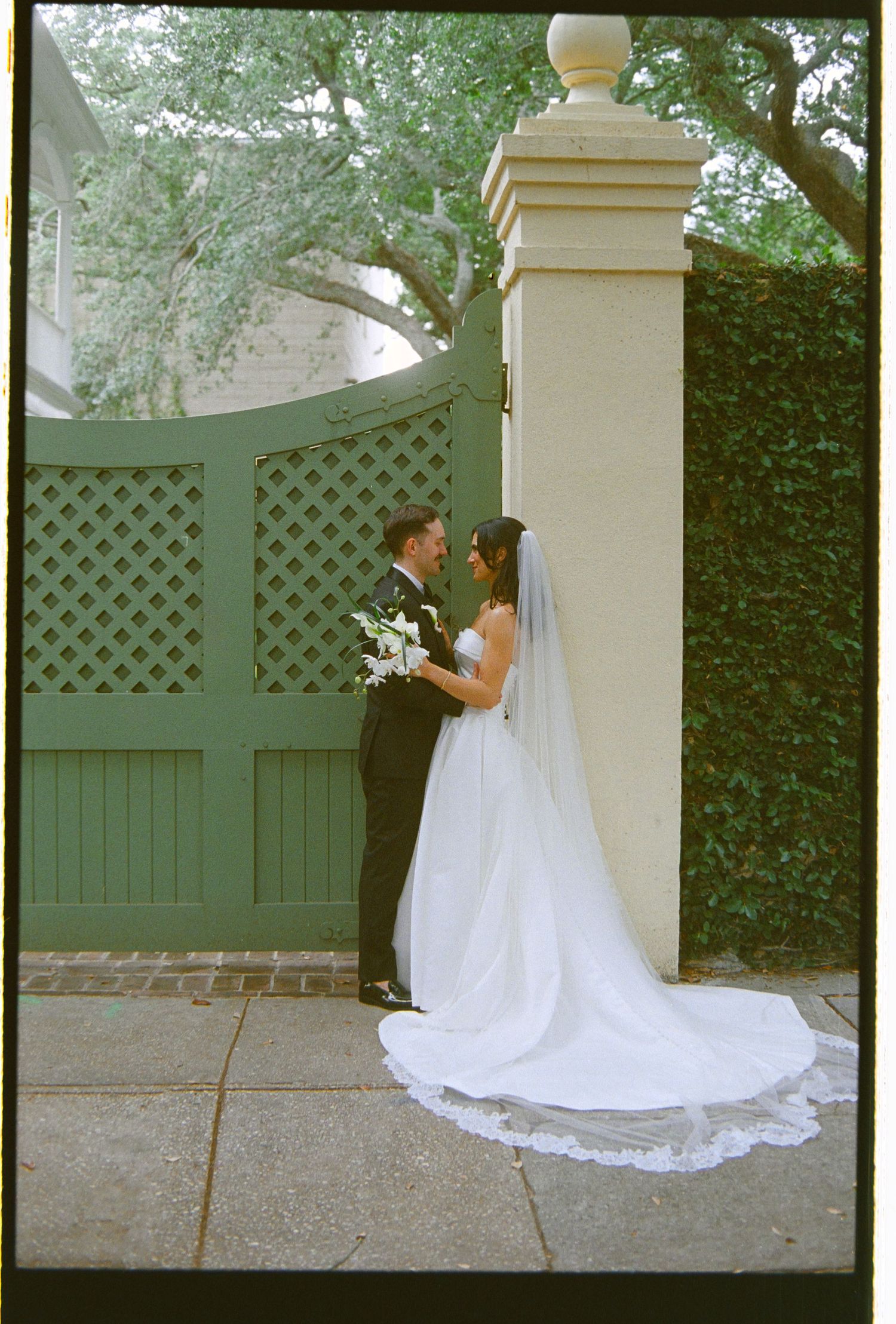 Bride and groom sharing intimate moment by ornate green garden gate on wedding day.