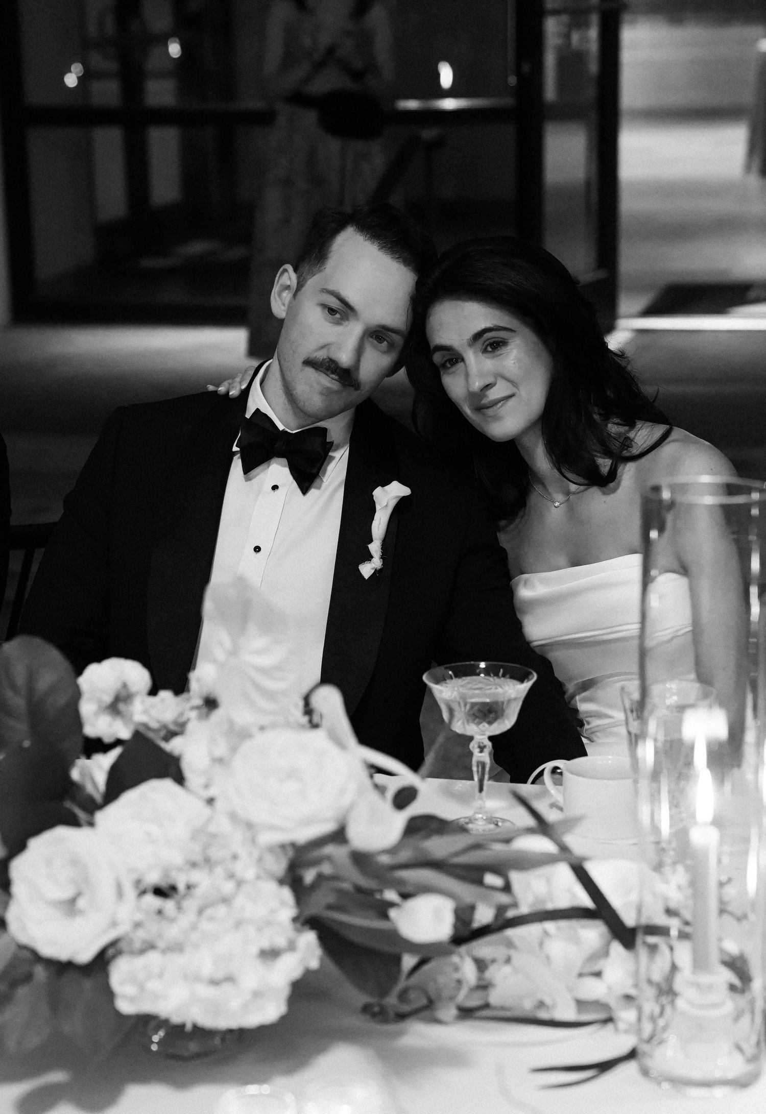 Black and white portrait of bride and groom seated together at wedding reception table.