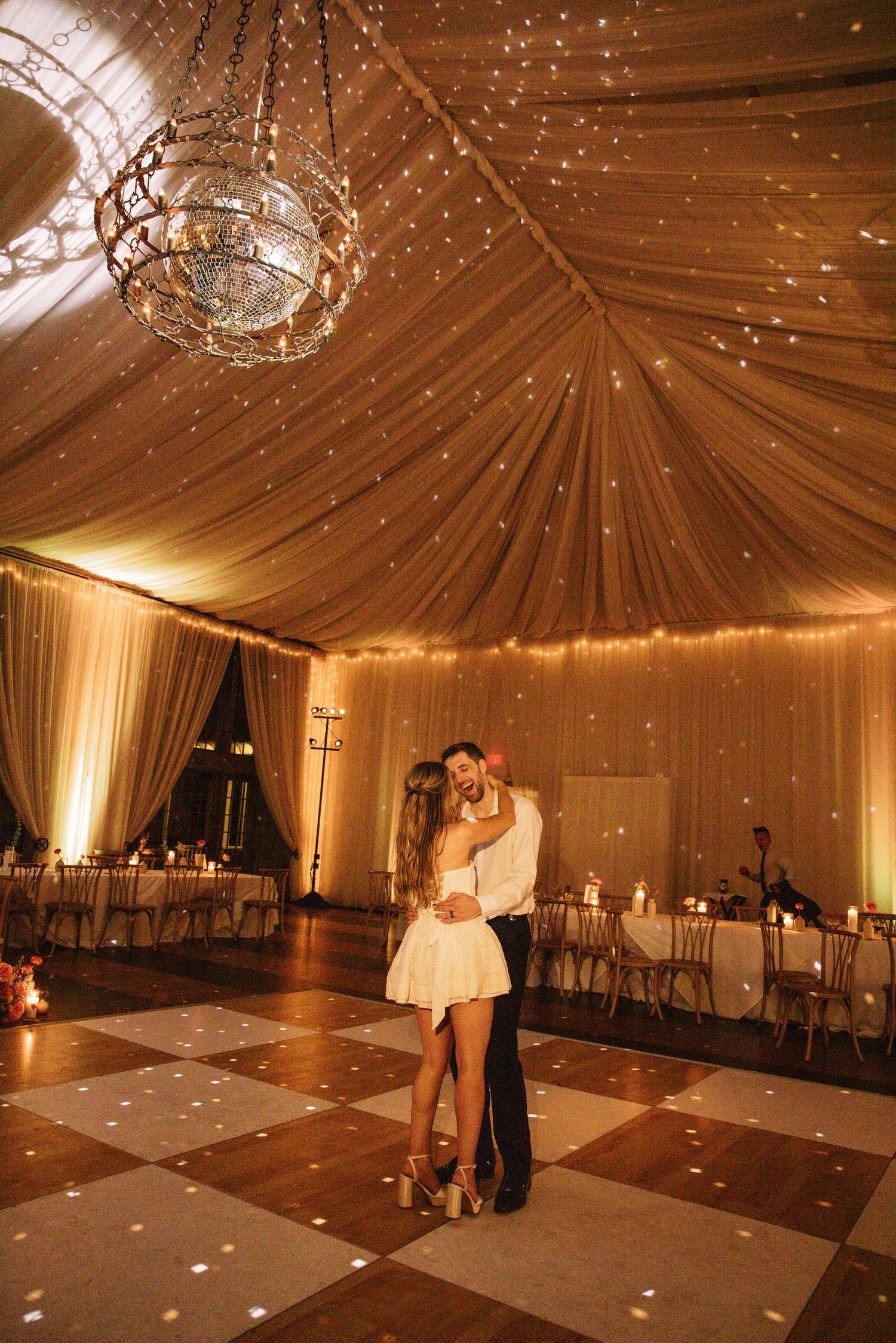 Bride and groom share first dance under glowing chandelier and twinkling fairy lights in draped tent.