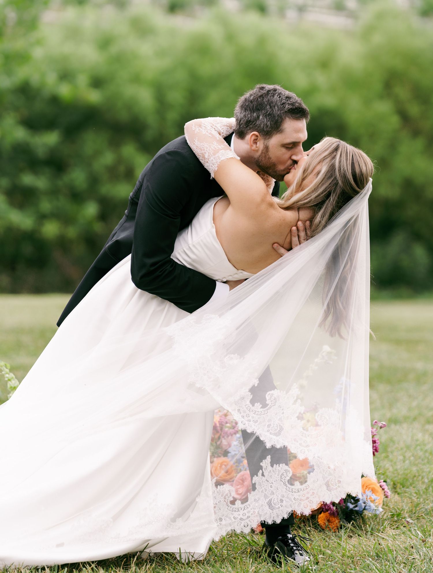 Bride and groom share a romantic dip kiss outdoors, her lace-trimmed veil flowing gracefully over green grass.