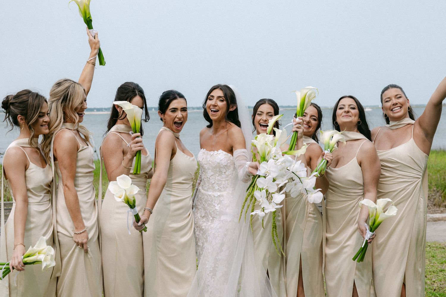 Wedding party in neutral dresses lines up for a celebratory photo by the water.