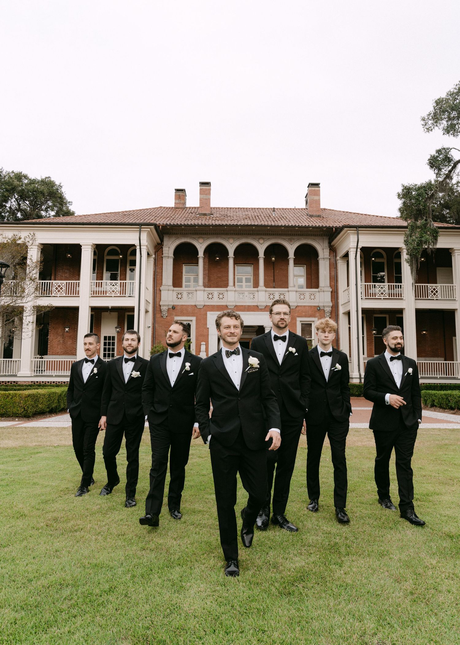 Groomsmen in black tuxedos walk together across a green lawn in front of a brick building with white columns.