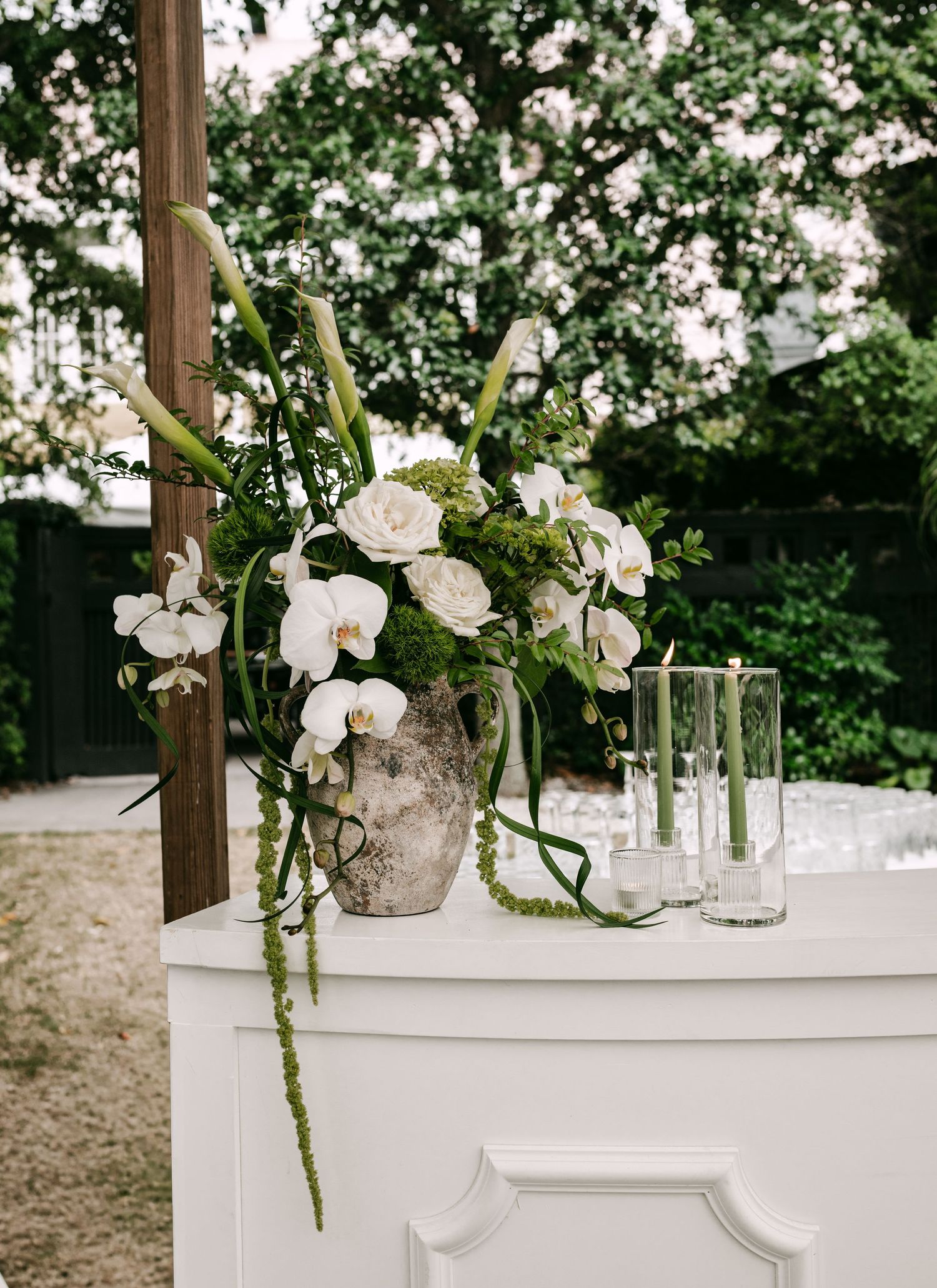 Elegant floral arrangement with white orchids and roses in a stone vase on a white cabinet at an outdoor wedding.