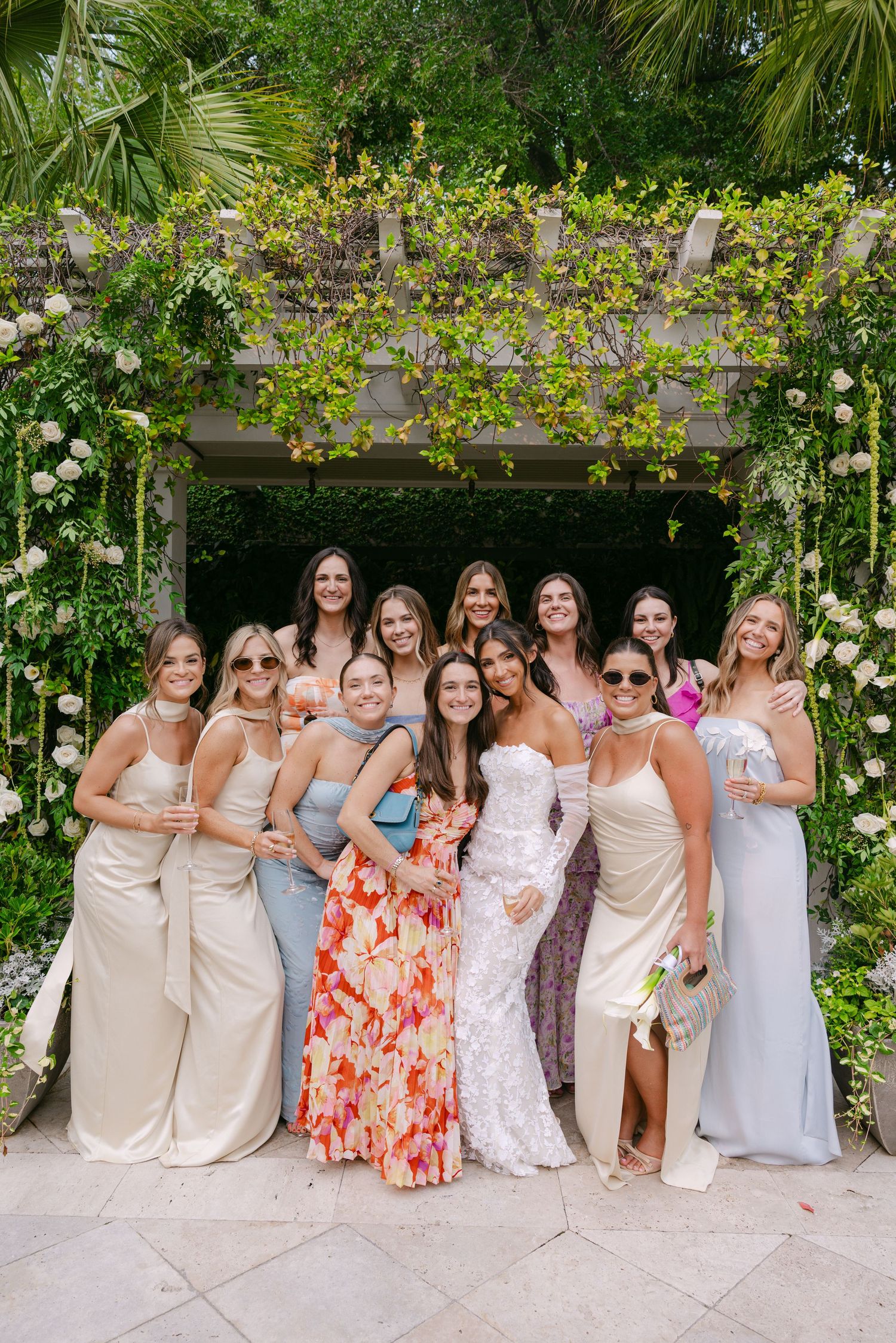 A group in formal attire poses together against a lush garden backdrop with trailing vines and greenery.