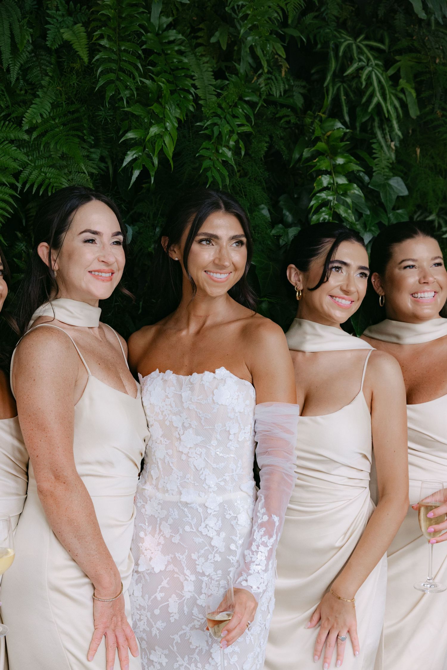 Line of bridesmaids in white and cream dresses pose together against a green foliage backdrop.