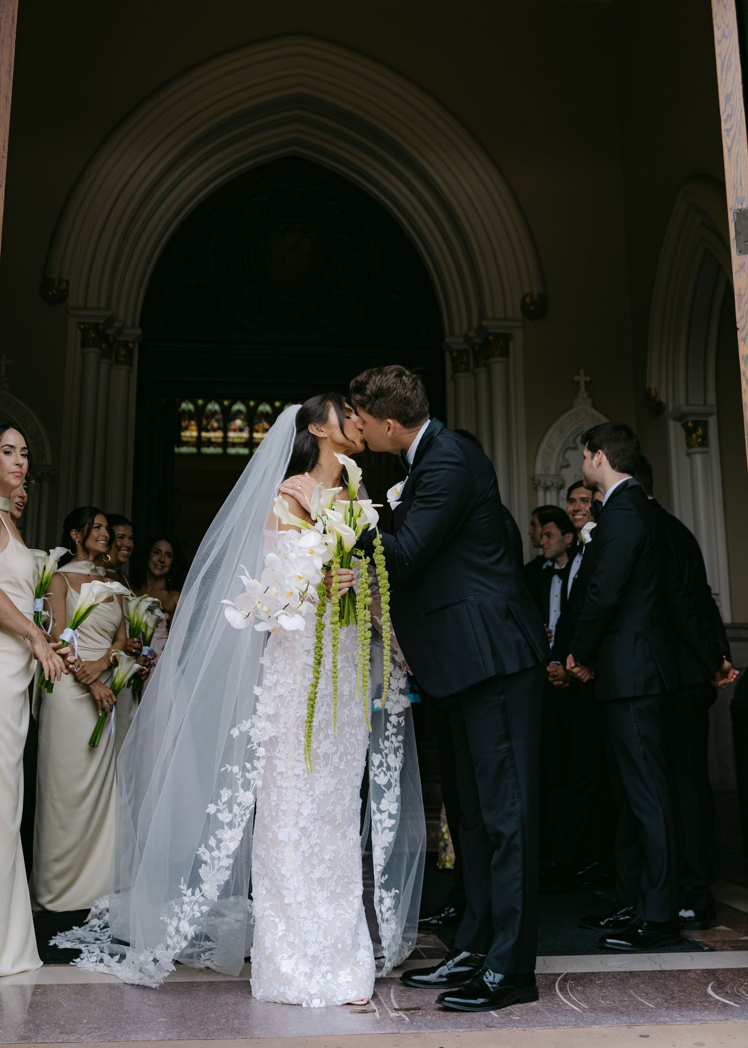 Wedding party gathers at church entrance with bride in white lace gown and bridesmaids in cream dresses.