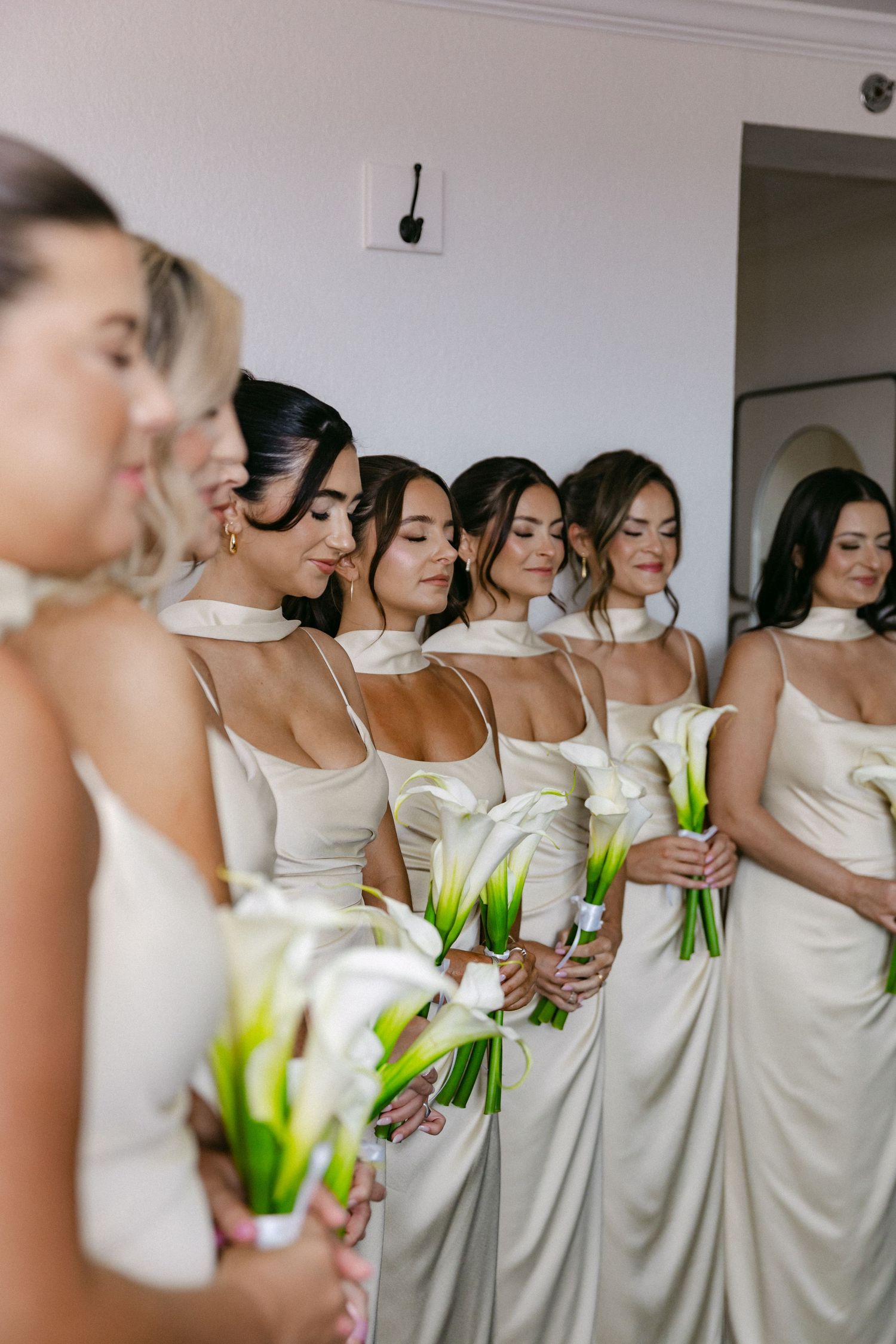 A row of bridesmaids in matching champagne satin dresses holding white calla lily bouquets.
