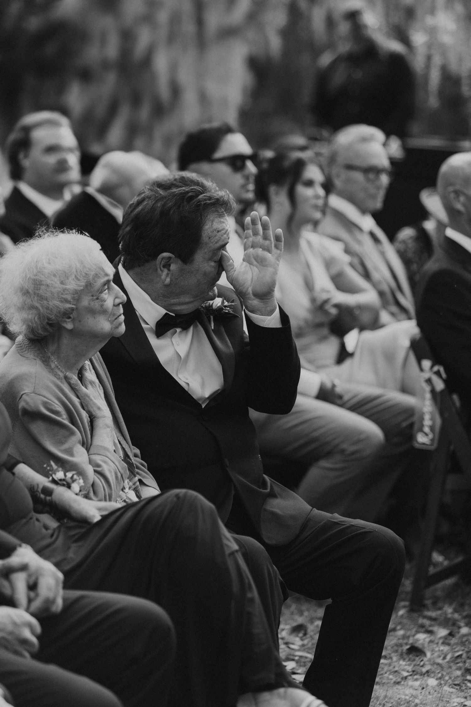 Wedding guests seated in formal attire watch the ceremony outdoors.