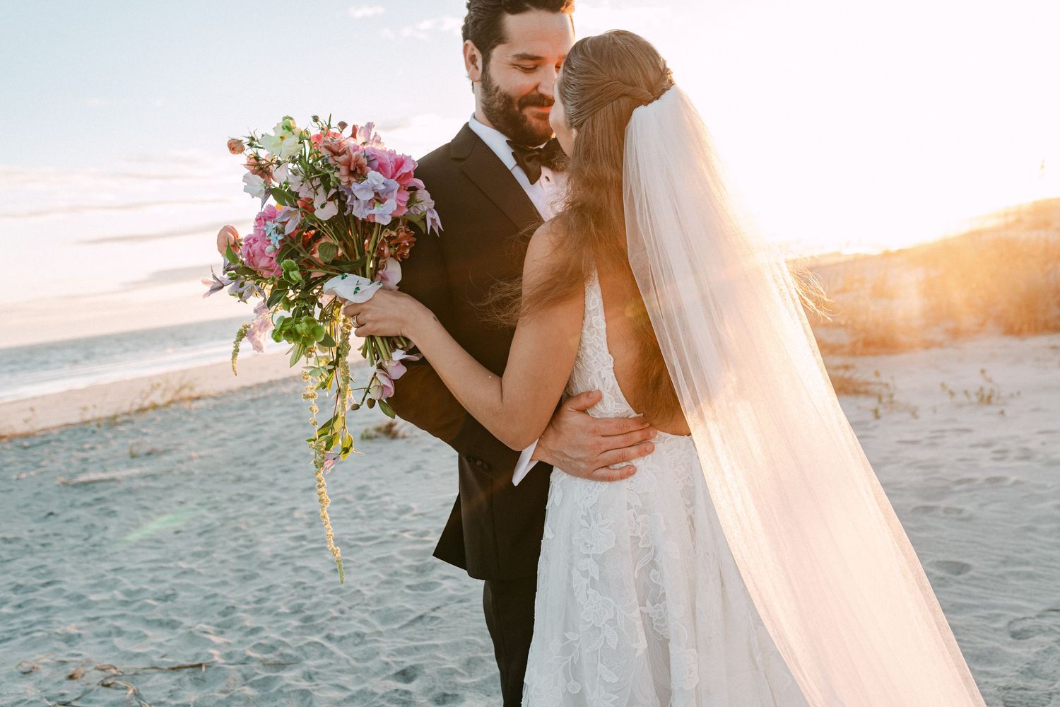 Bride and groom embracing on beach at golden sunset, bride holding vibrant pink floral bouquet.