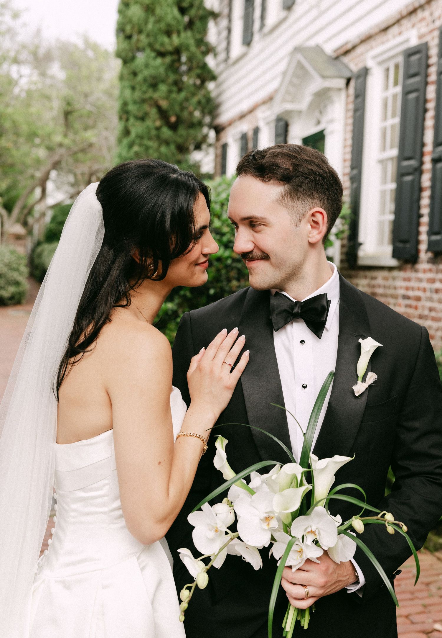 Bride and groom share an intimate moment outside a brick building, groom holding white orchid bouquet.