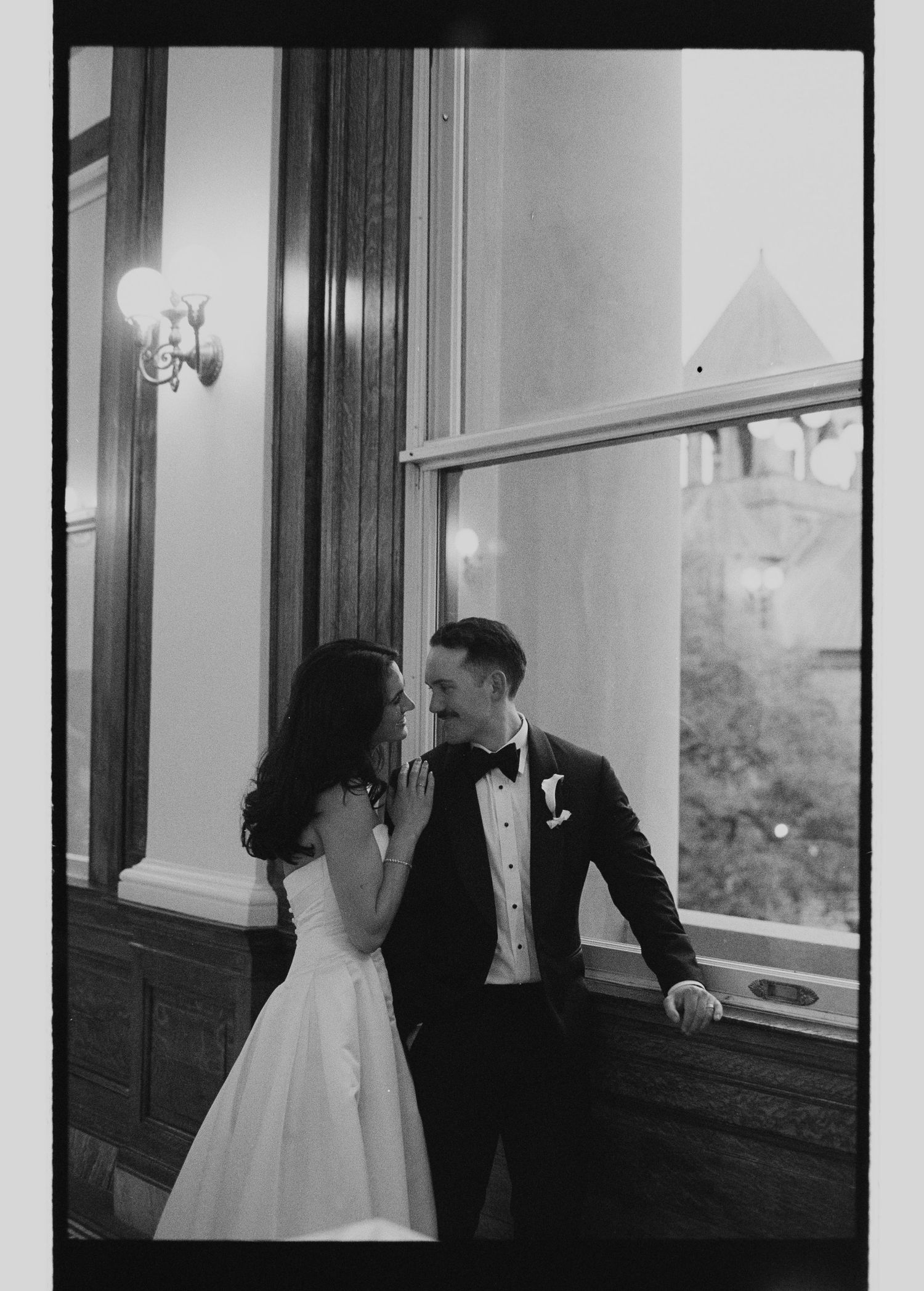 Black and white photo of bride and groom sharing an intimate moment by a large window indoors.