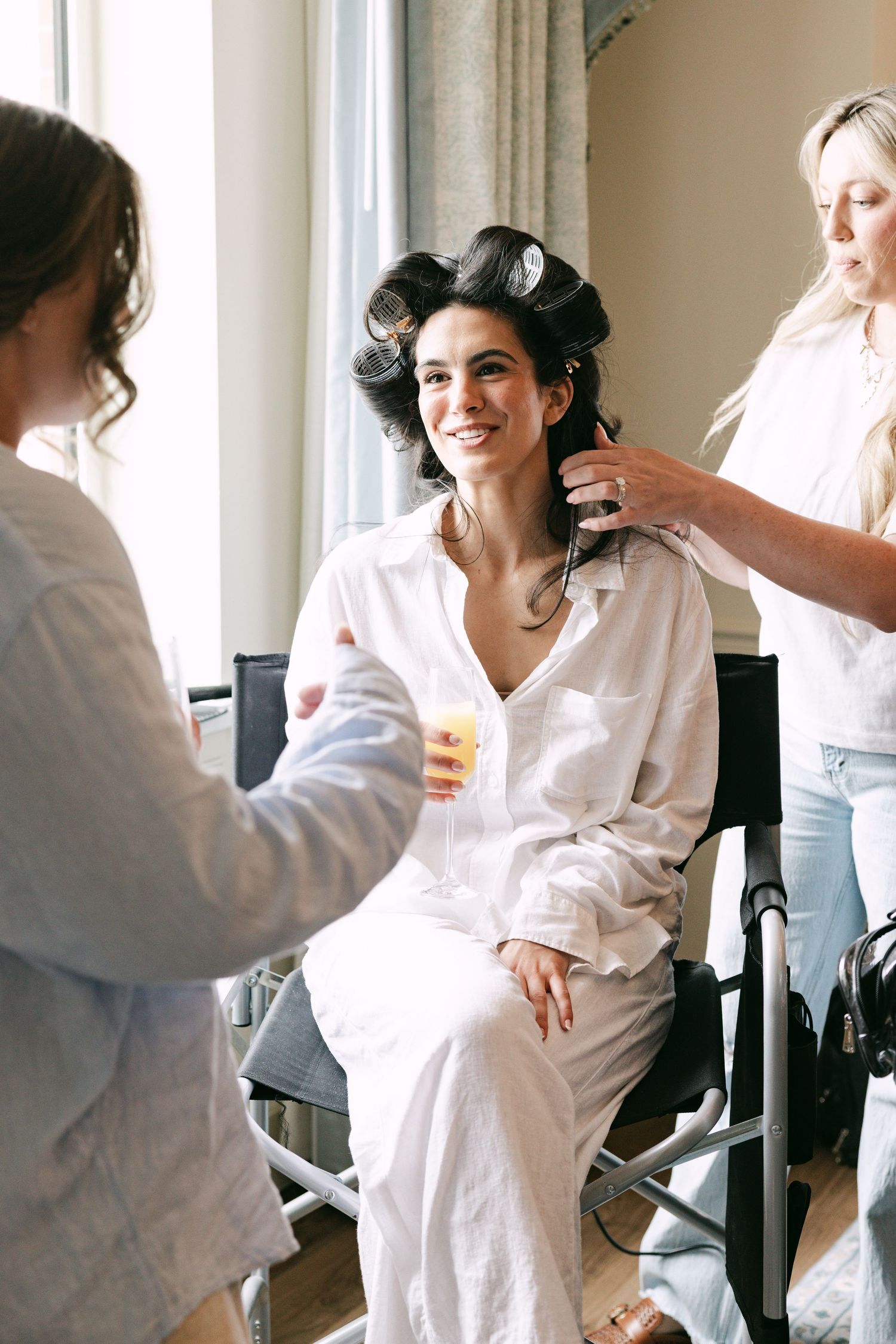 Smiling bride in white robe with hair rollers gets styled by hairdresser while bridesmaid offers a drink.