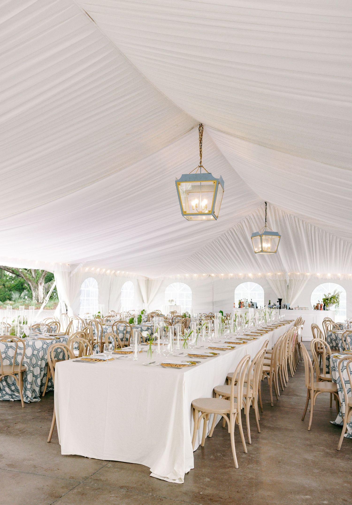 Spacious white wedding tent reception with long tables, bentwood chairs, floral linens, and hanging lantern centerpieces.
