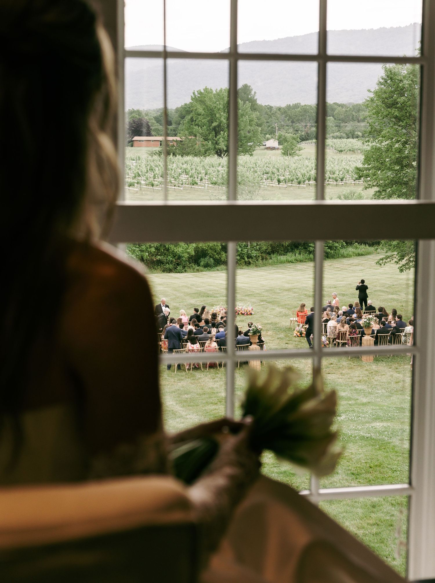 Bride holding bouquet gazes through window at outdoor wedding ceremony on lush green lawn with mountain views.