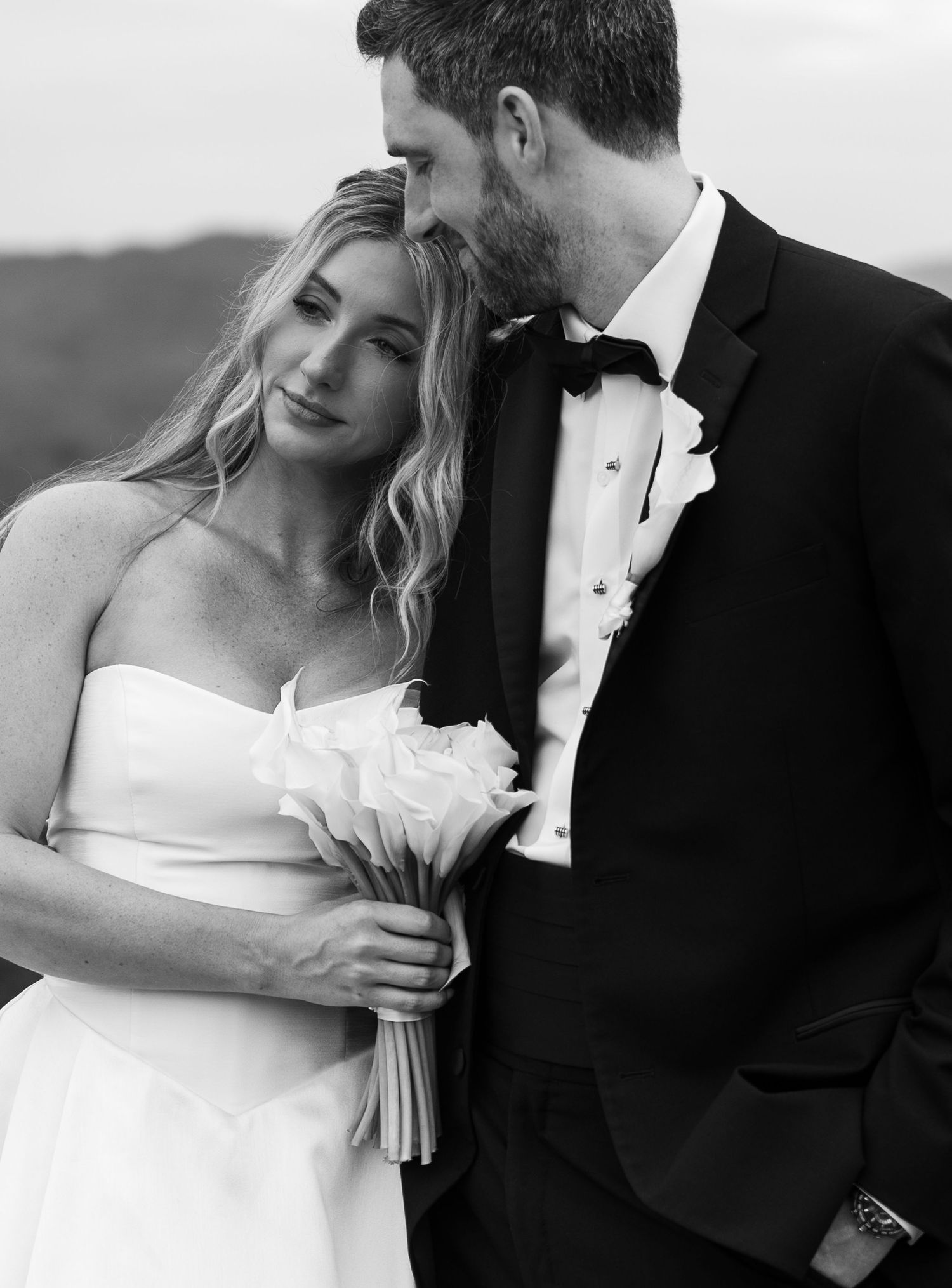 Black and white wedding photo of bride holding white bouquet leaning close to groom in tuxedo outdoors.