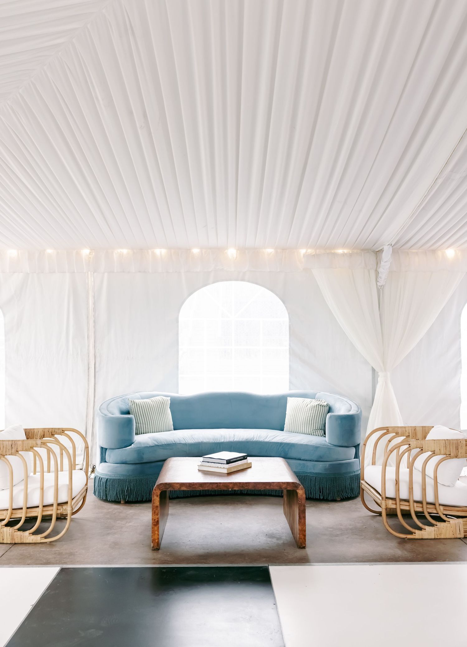 Elegant tent lounge with blue velvet curved sofa, rattan chairs, wooden coffee table, and white draped ceiling with string lights.