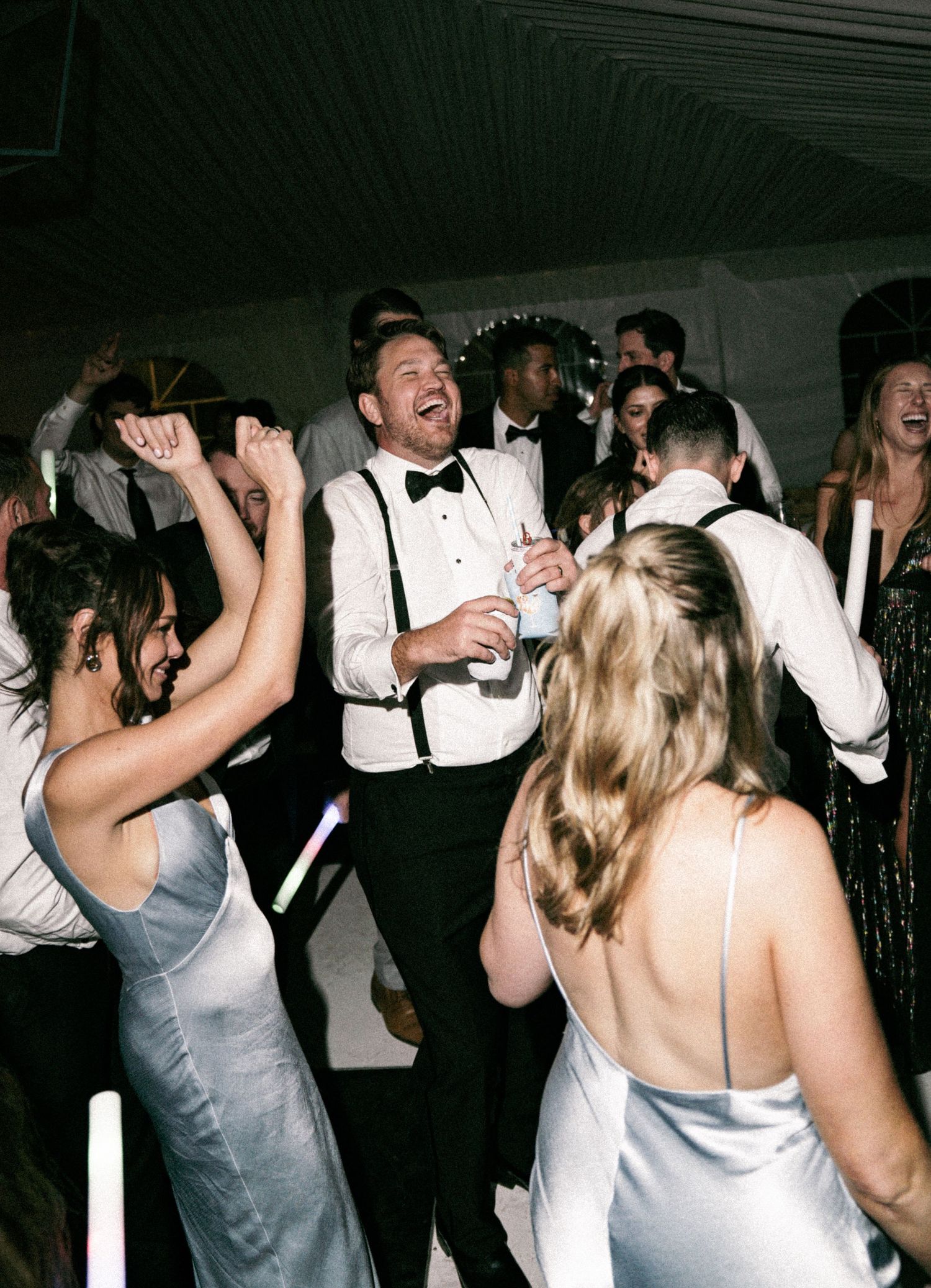 Guests in formal attire dancing joyfully at a wedding reception, with the groom laughing in a tuxedo and suspenders.