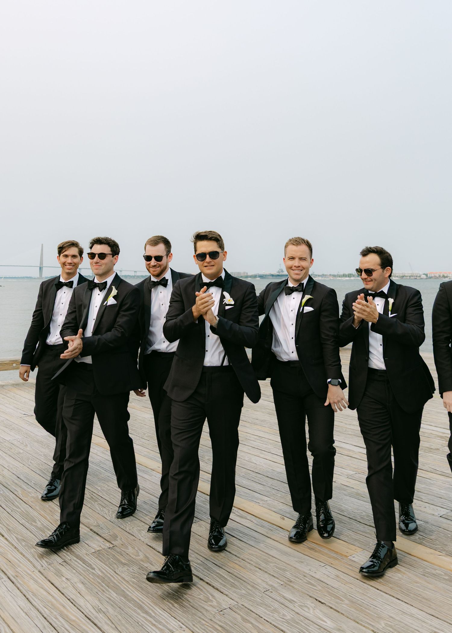 Wedding party groomsmen in black suits posing casually on oceanside deck.