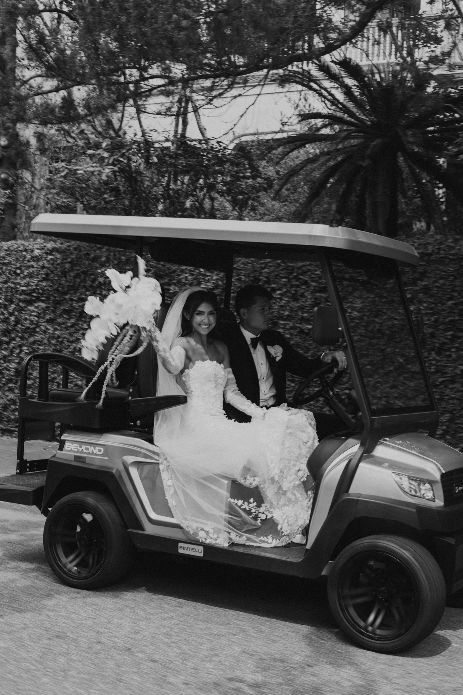 Wedding couple riding in decorated black golf cart through tropical setting.
