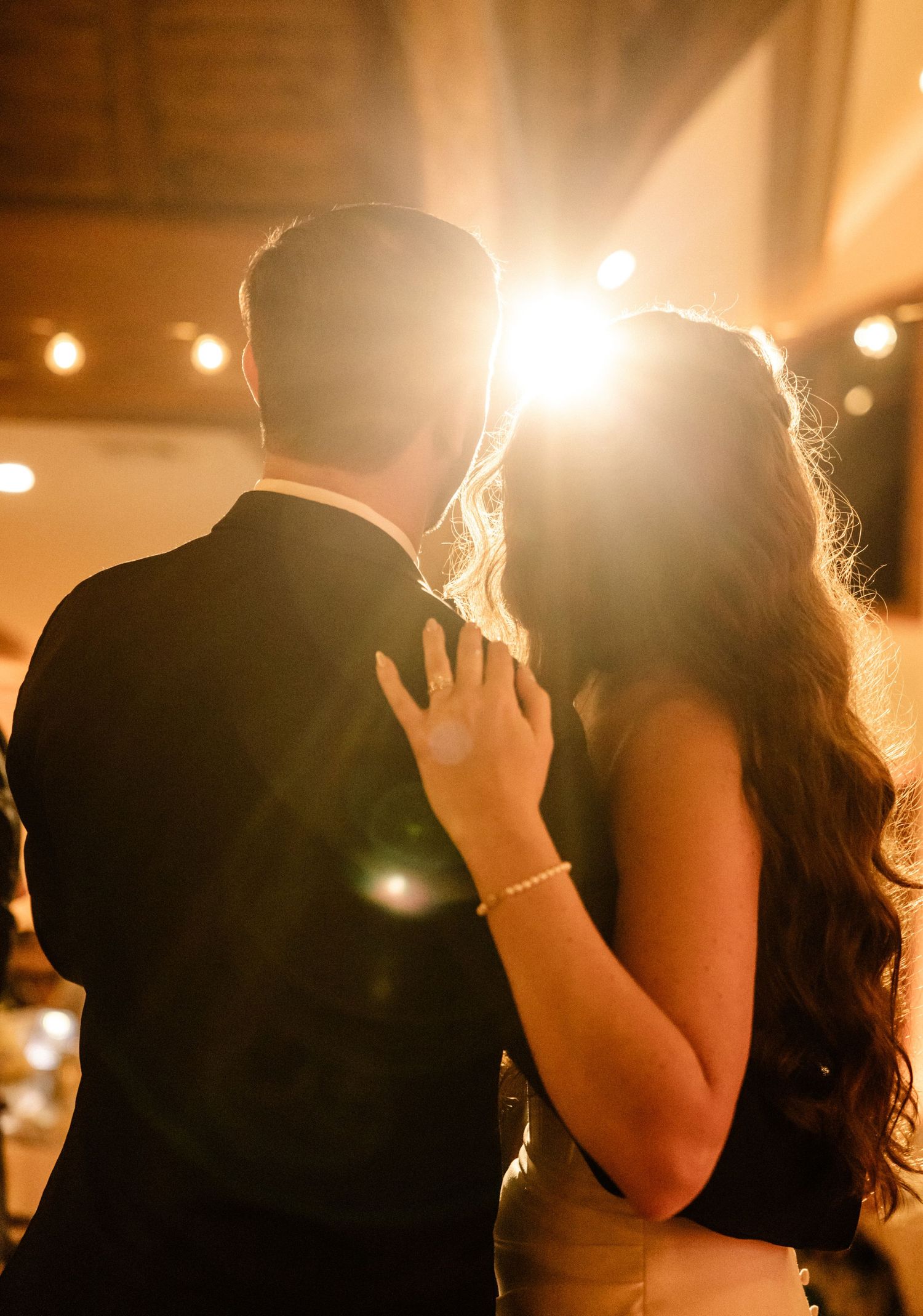 Couple sharing a romantic first dance kiss backlit by warm golden light at an elegant wedding reception.