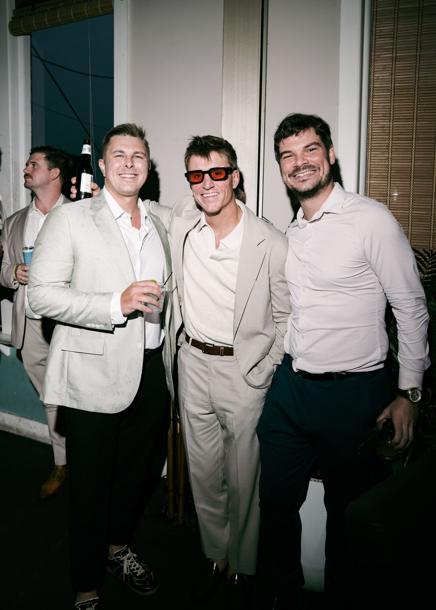 Three stylish men in neutral toned outfits smiling together at an upscale social event.