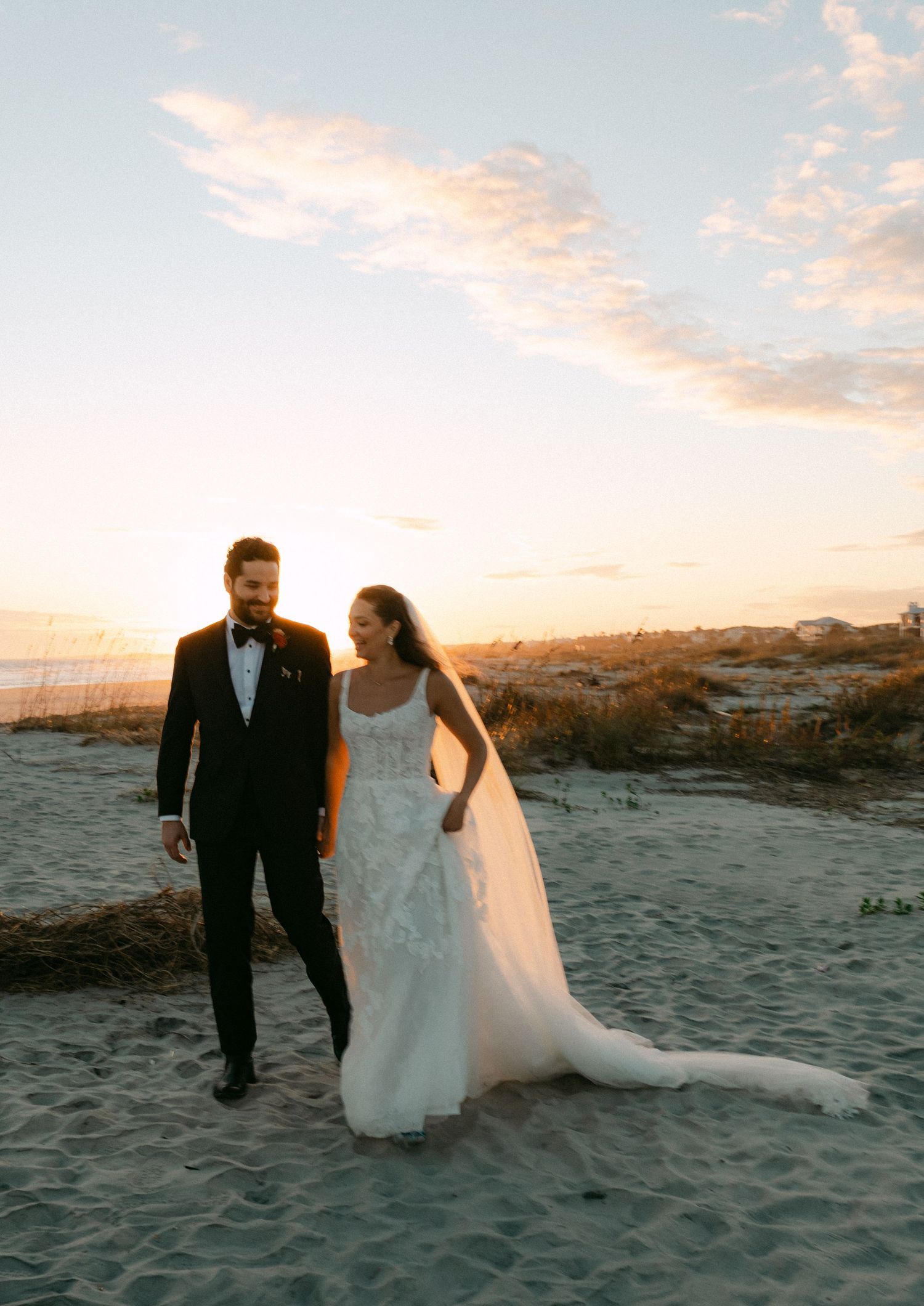 Bride and groom walking on sandy beach at sunset, bride in lace gown with flowing veil.