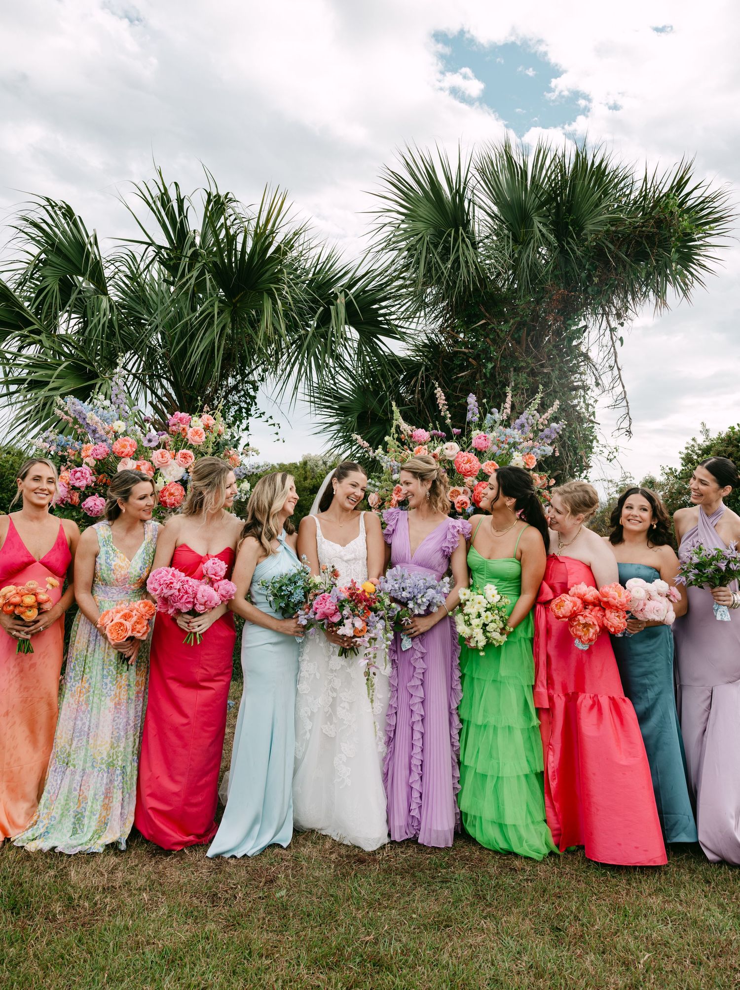 Bride surrounded by bridesmaids in vibrant multicolored gowns holding bright floral bouquets outdoors.