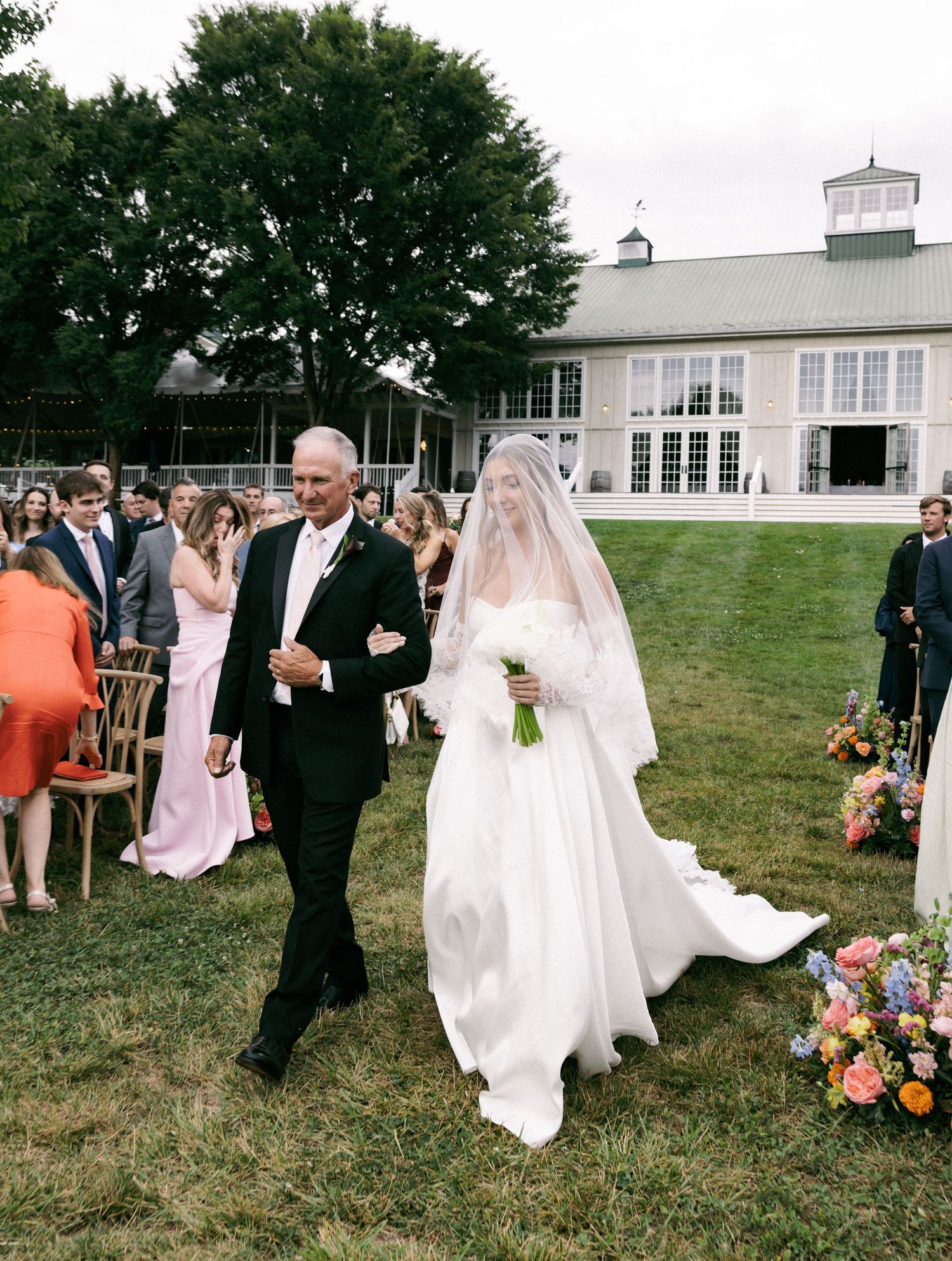 Bride in white gown and veil walks down outdoor aisle with escort at elegant garden wedding ceremony.