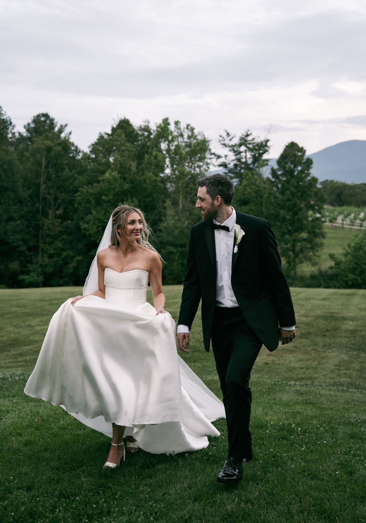 Bride and groom walking on green lawn, she in white ballgown, he in dark tuxedo, with mountain views.
