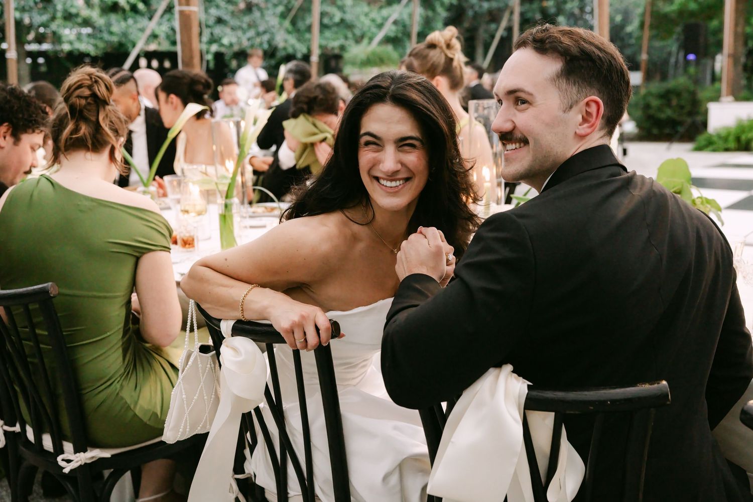 Smiling bride in strapless gown laughing with groom in black suit at outdoor wedding reception.
