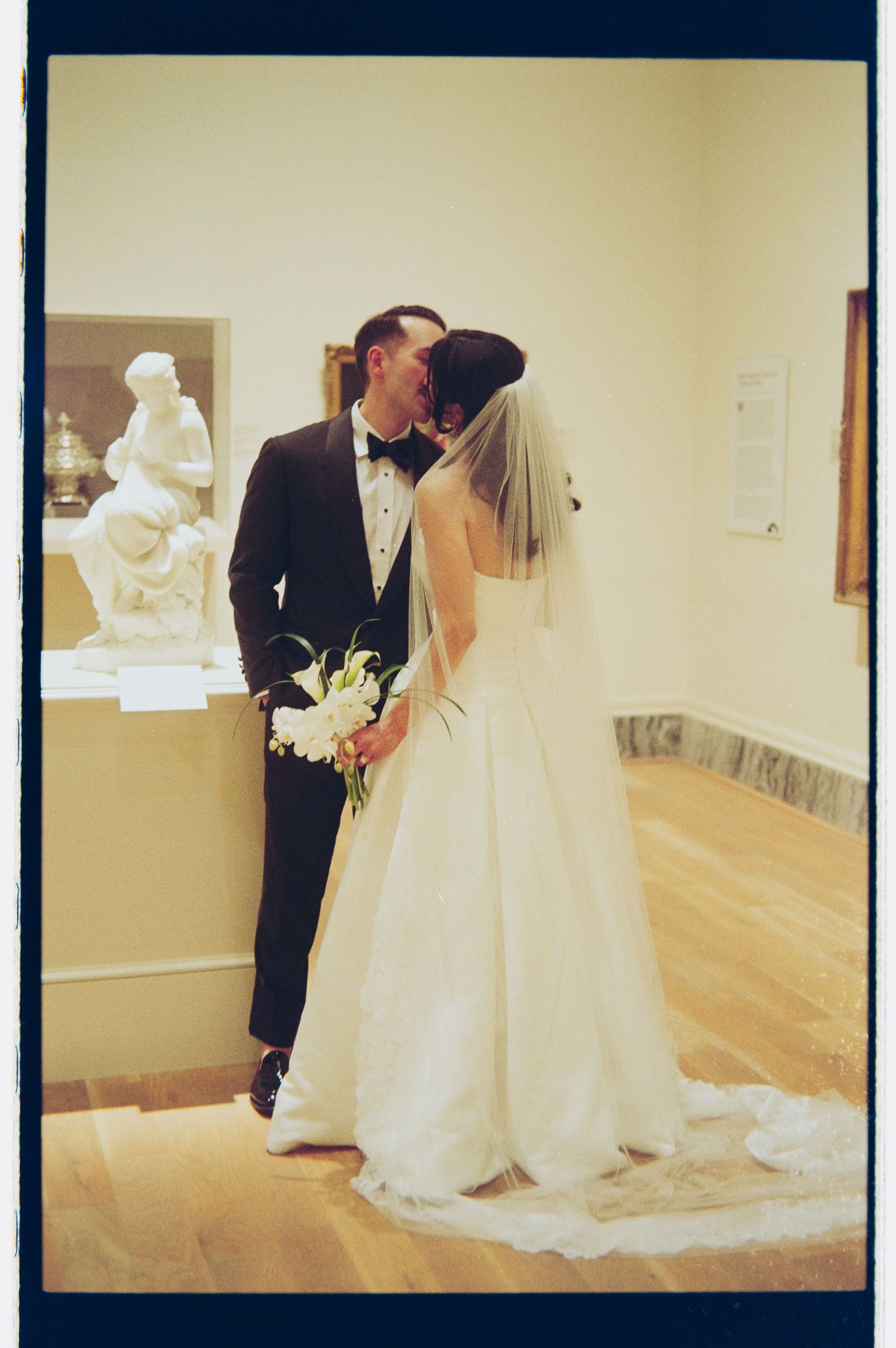 Bride and groom sharing a kiss in an art museum gallery, bride holding white floral bouquet with veil.