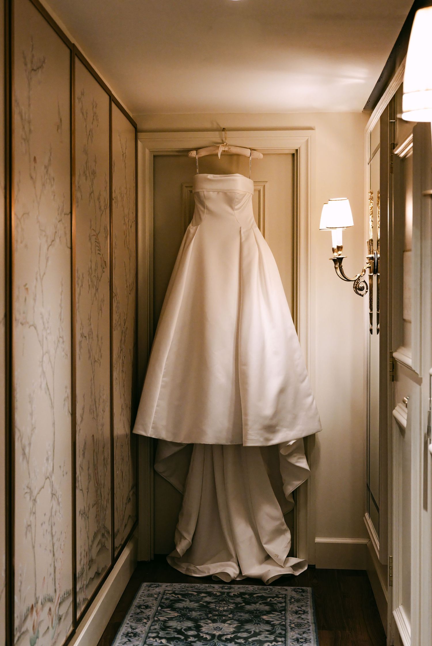 A strapless white ball gown wedding dress hanging in an elegant hallway with warm wall sconce lighting.