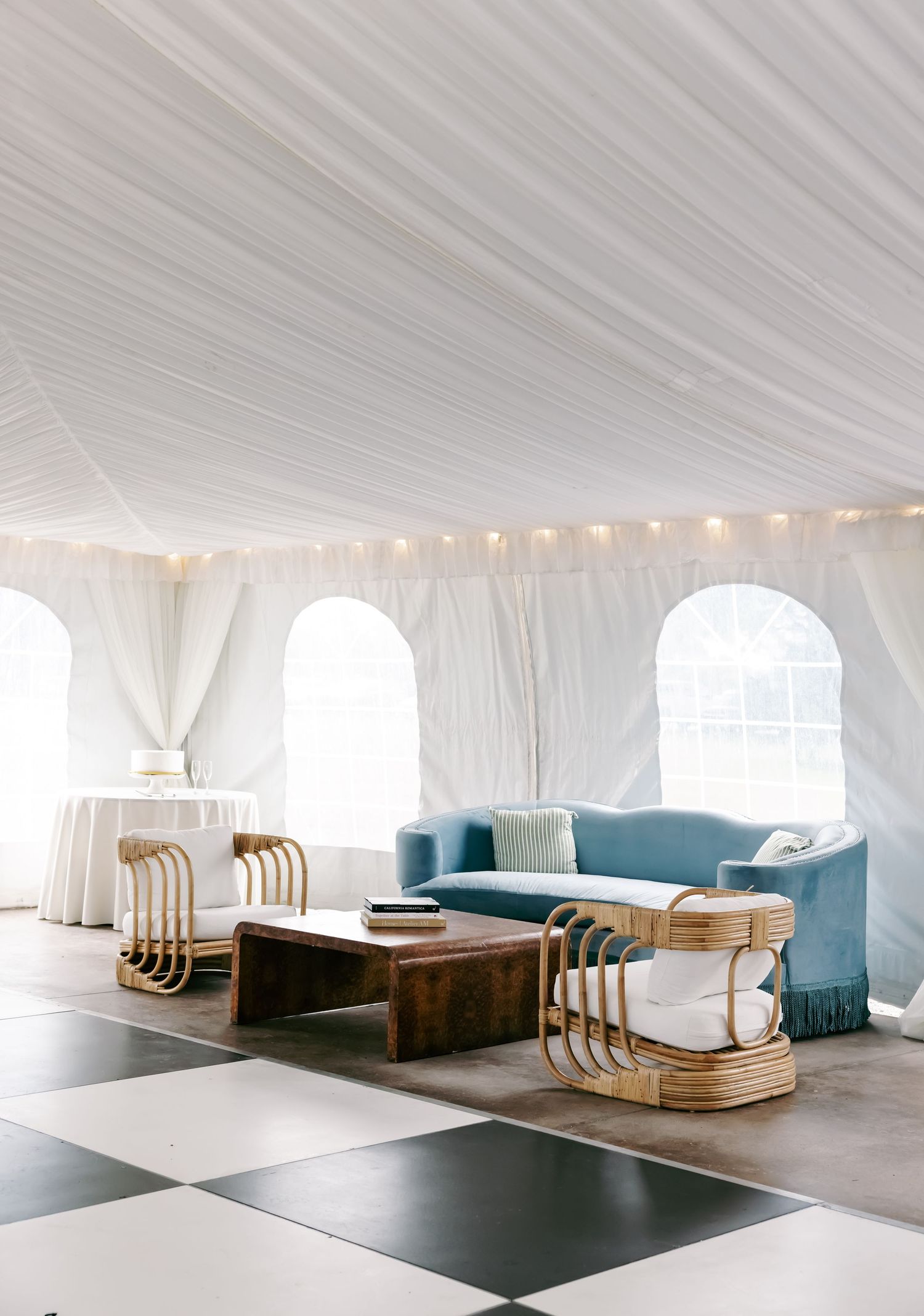 Elegant wedding tent reception lounge with blue velvet sofa, rattan chairs, and checkered floor.