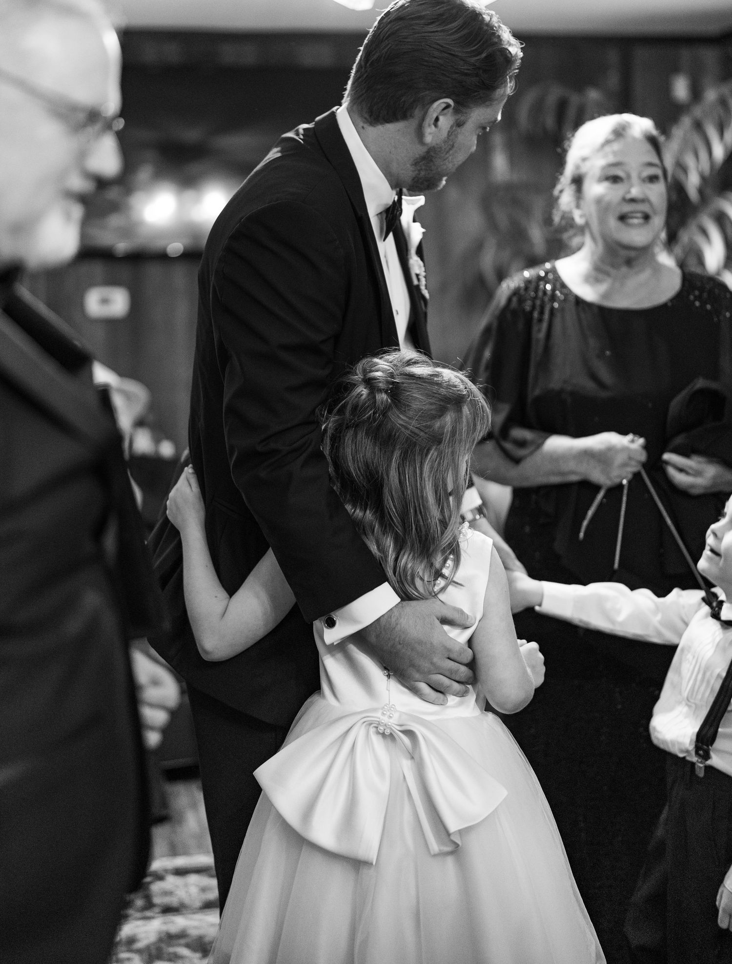Groom in tuxedo hugging flower girl in white dress with bow at wedding reception, black and white photo.