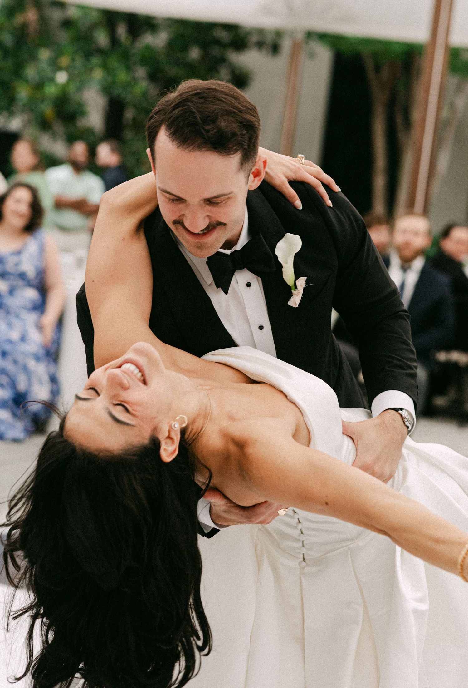 Groom dipping bride during first dance at wedding reception, both laughing joyfully in elegant attire.