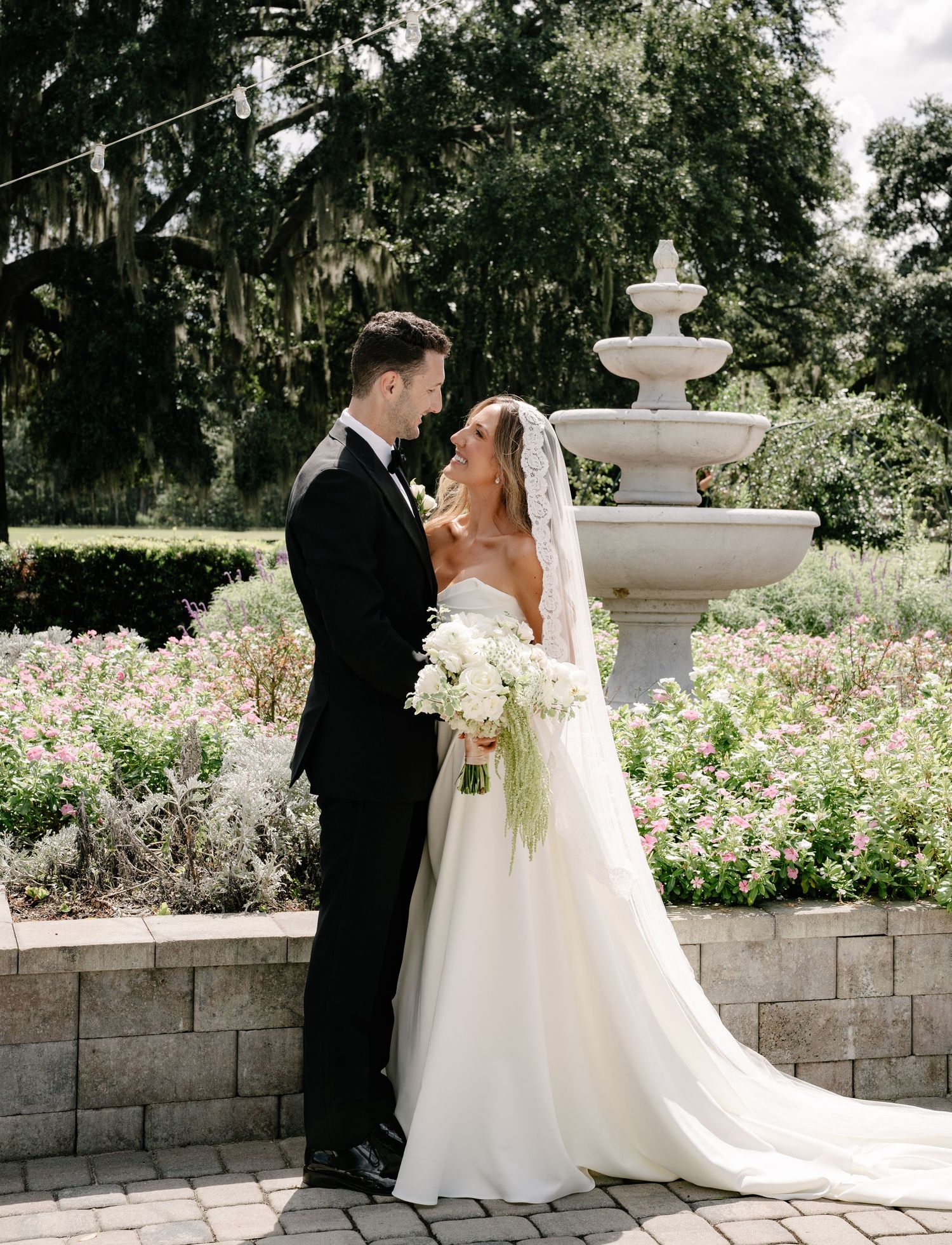 A couple shares a romantic moment in a garden setting with a decorative white fountain and blooming flowers.