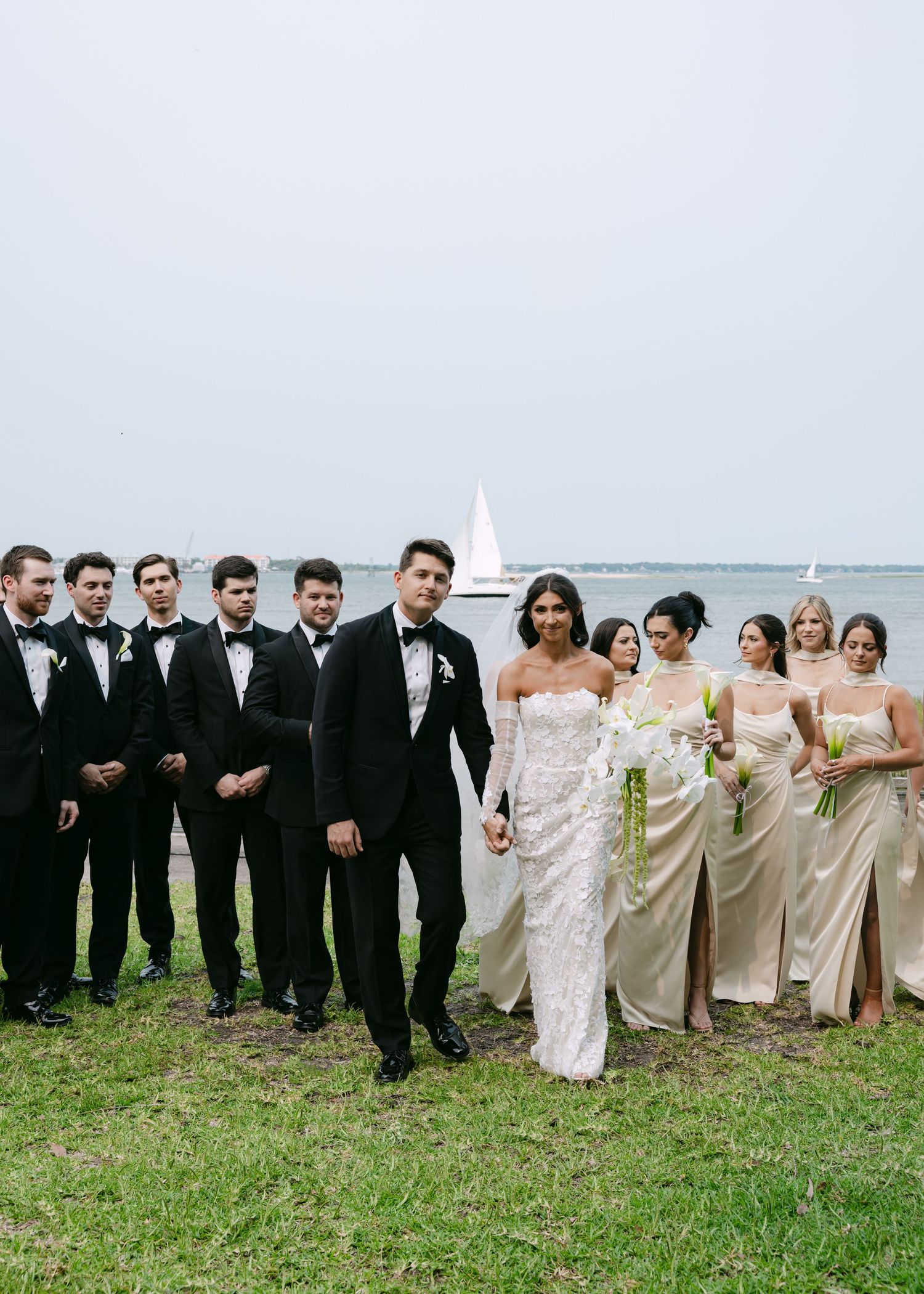 Full wedding party in formal attire poses together on grass with ocean backdrop.