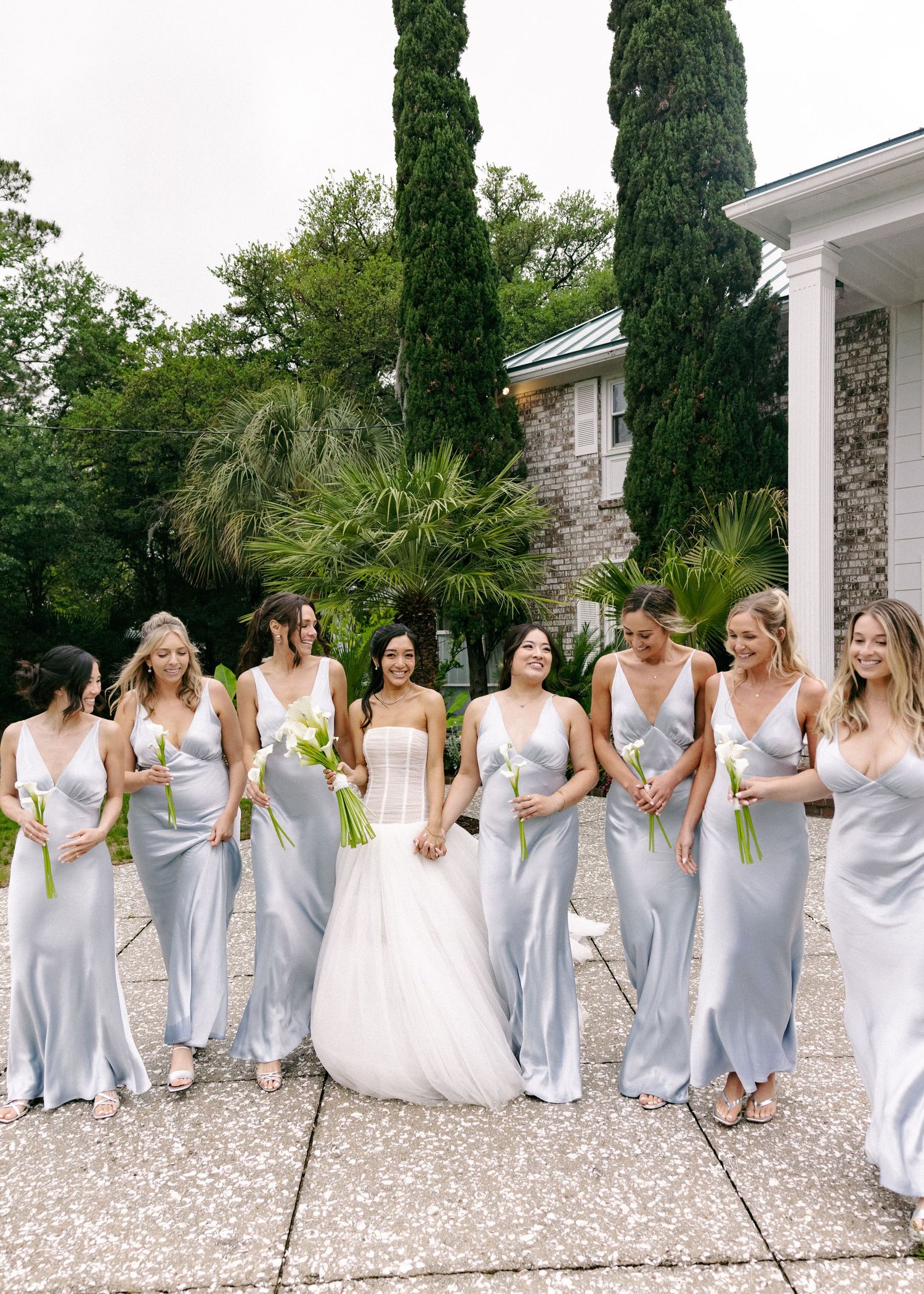 Bride in white corset gown poses with bridesmaids in silver satin dresses holding calla lilies outdoors.
