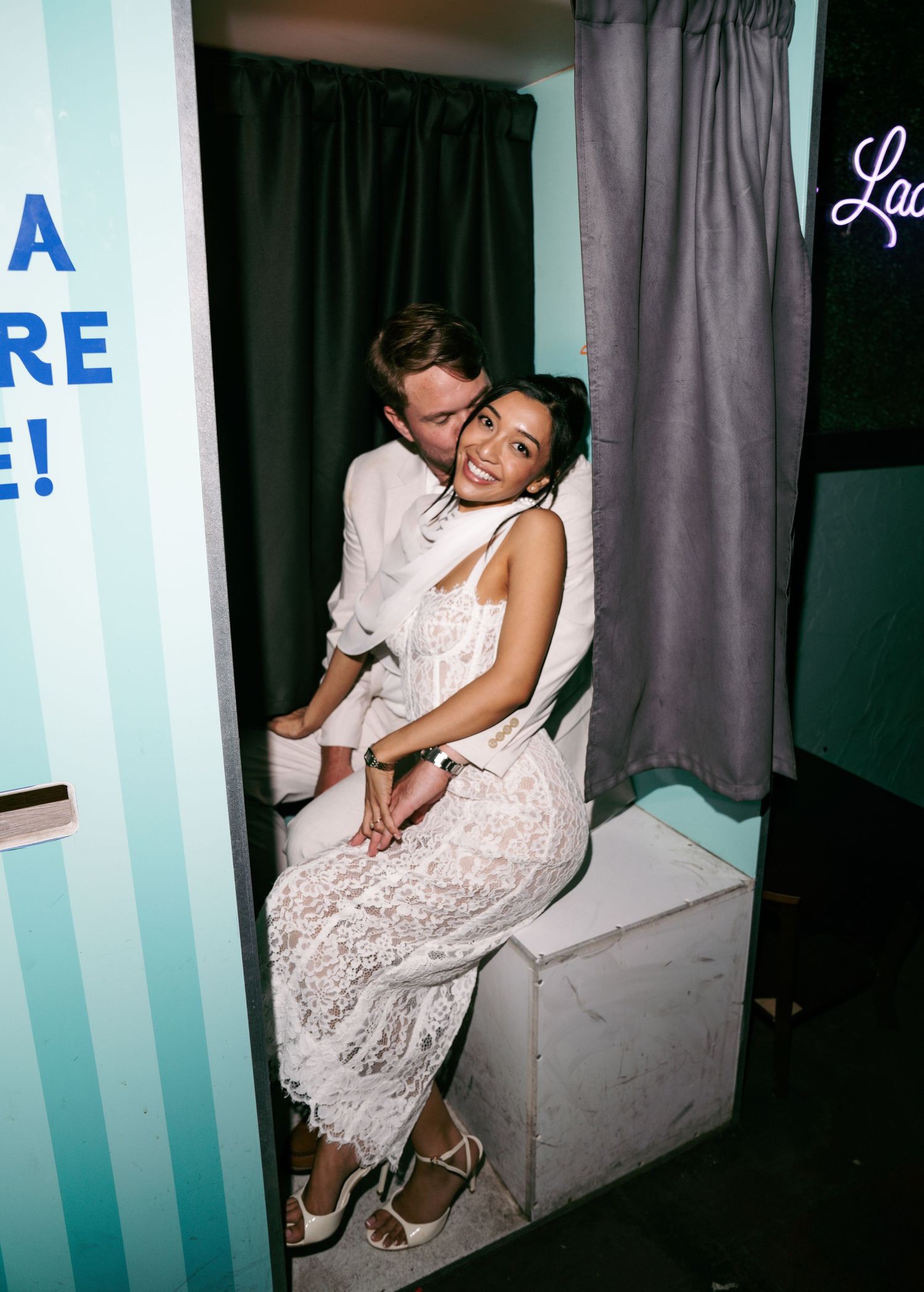 Couple posing together in a photo booth, woman in white lace dress and man in white shirt, smiling intimately.