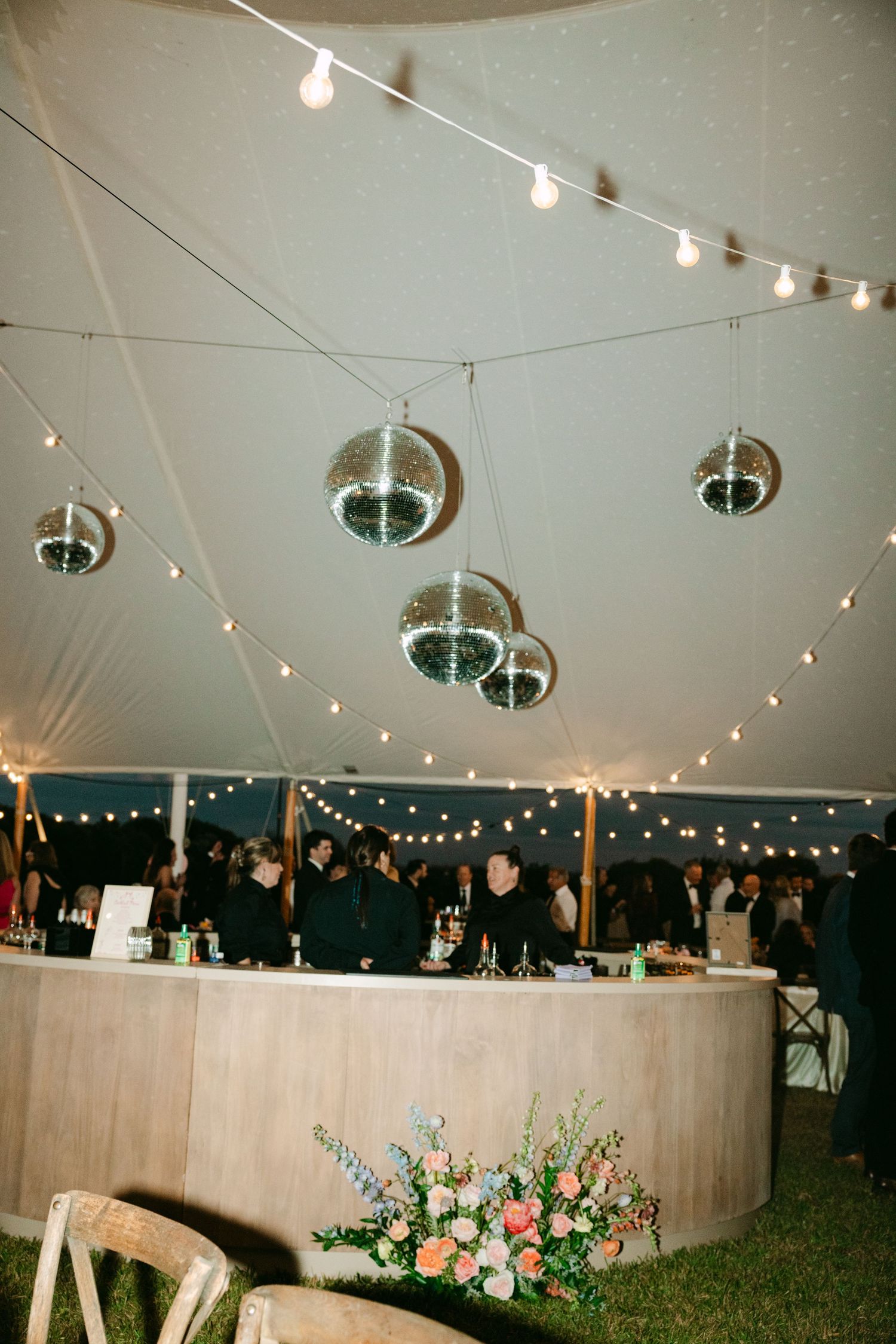 Wedding reception tent with disco balls, string lights, circular bar, and floral arrangements.