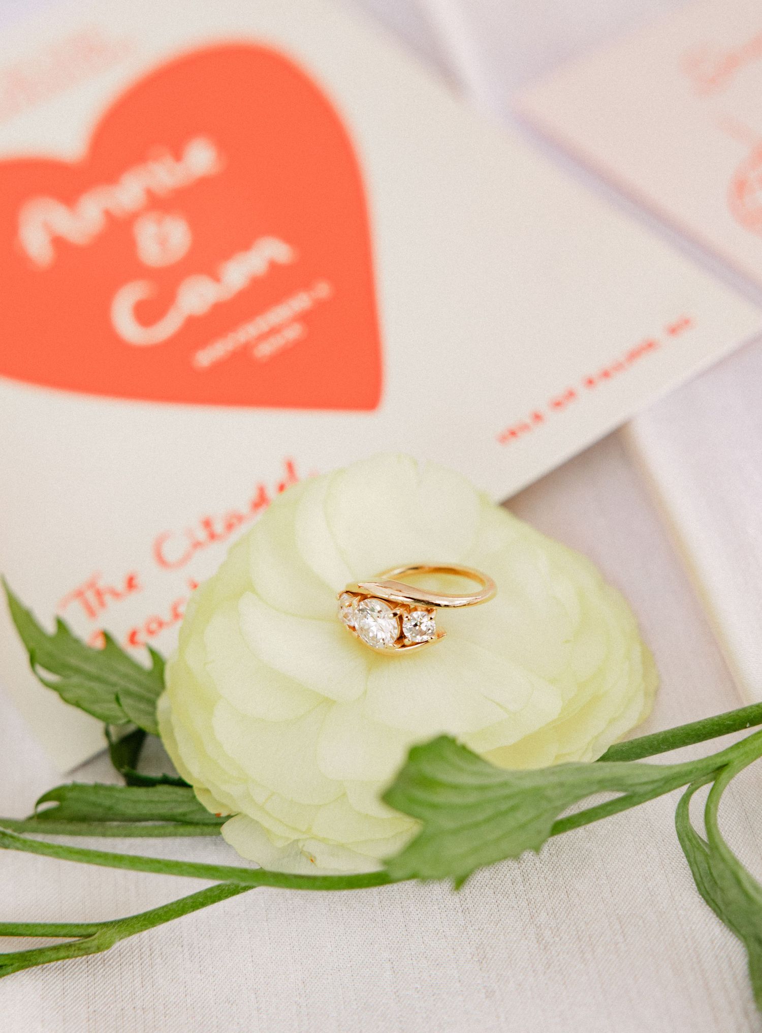 Gold diamond engagement ring rests on white ranunculus flower beside Annie and Cam wedding program.